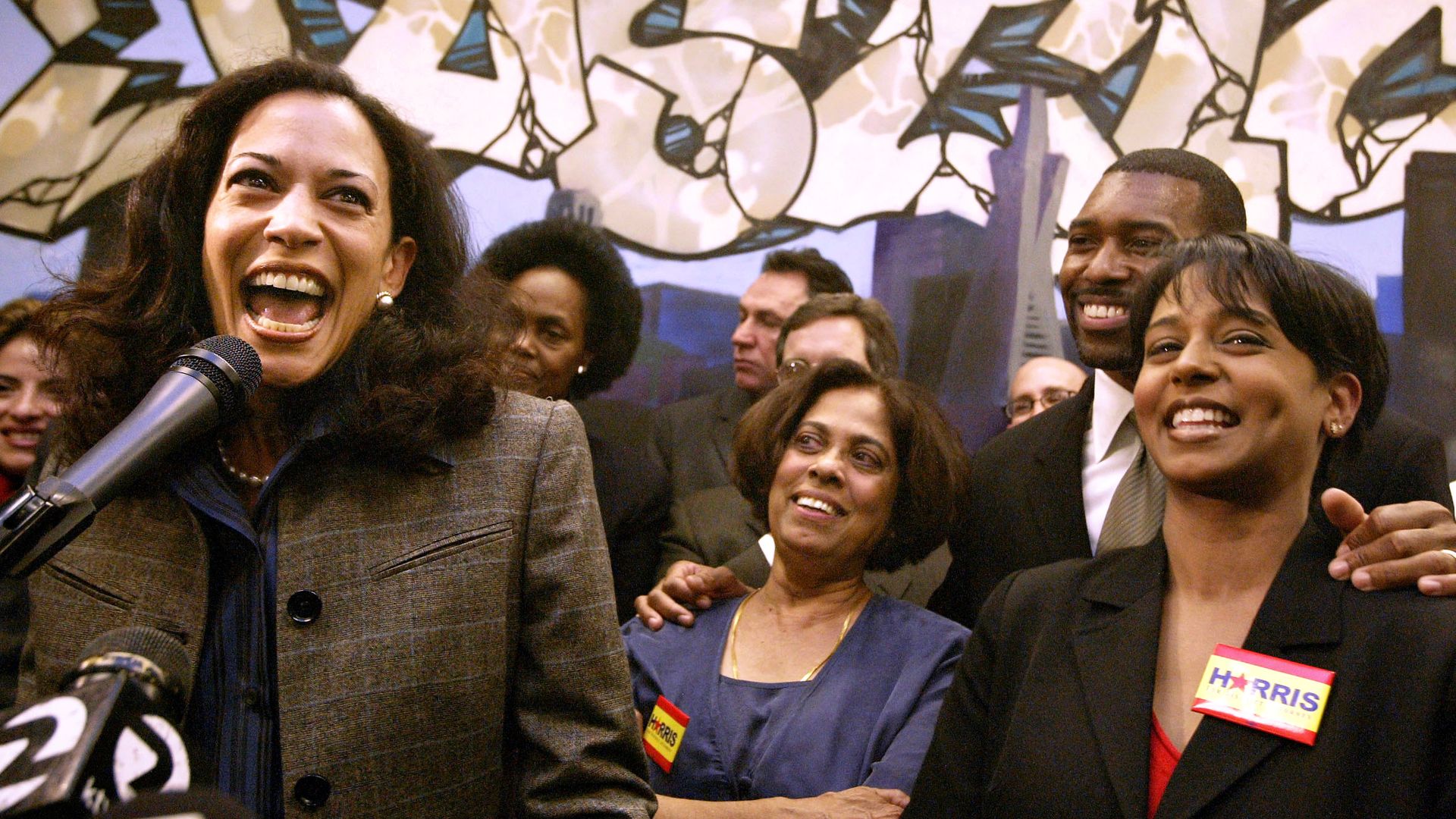 Photo of Kamala Harris speaking into a microphone as her mother and sister watch and smile from behind