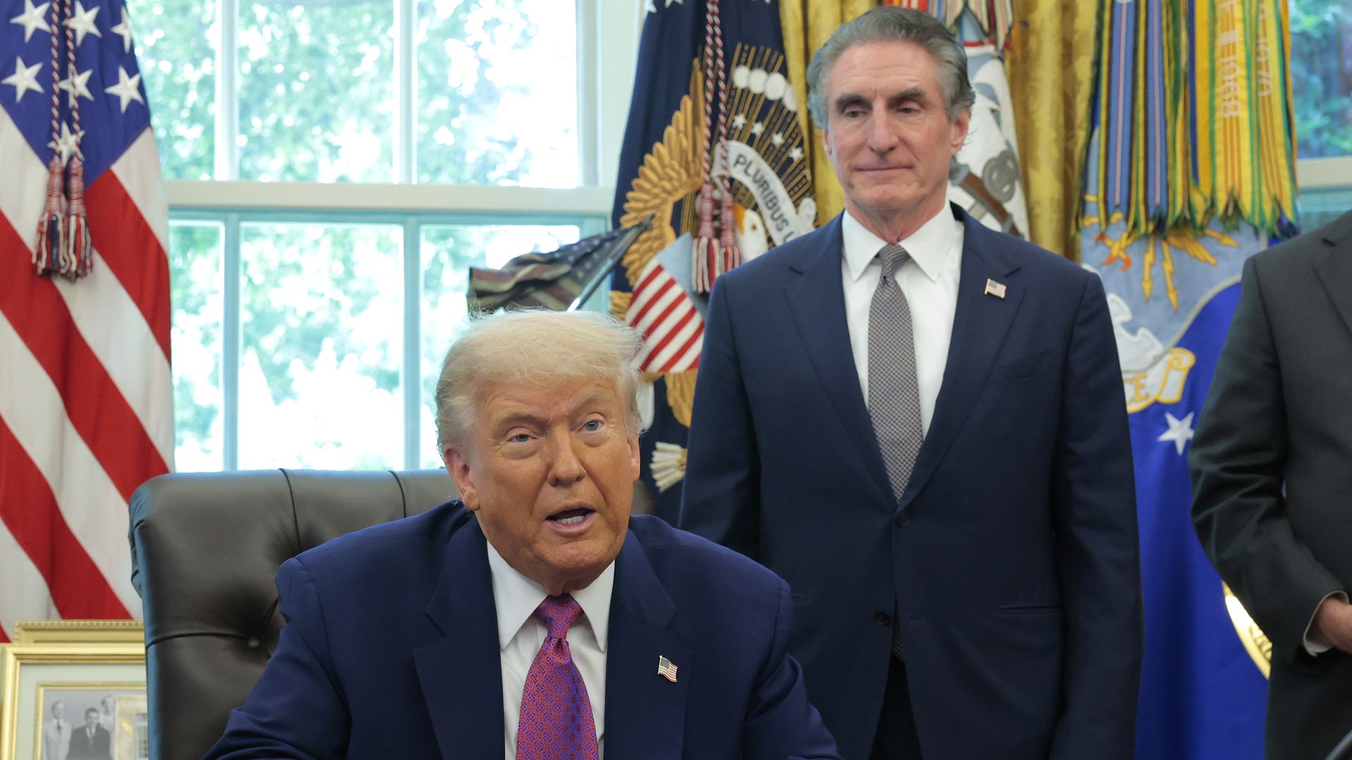 President Trump, wearing a navy jacket with a US flag pin at the top of his left lapel, white shirt and red tie, speaks as he sits at a brown desk while sitting in a brown leather chair as Interior Secretary Doug Burgum (R) wearing a navy jacket, white shirt and gray ties looks on.
