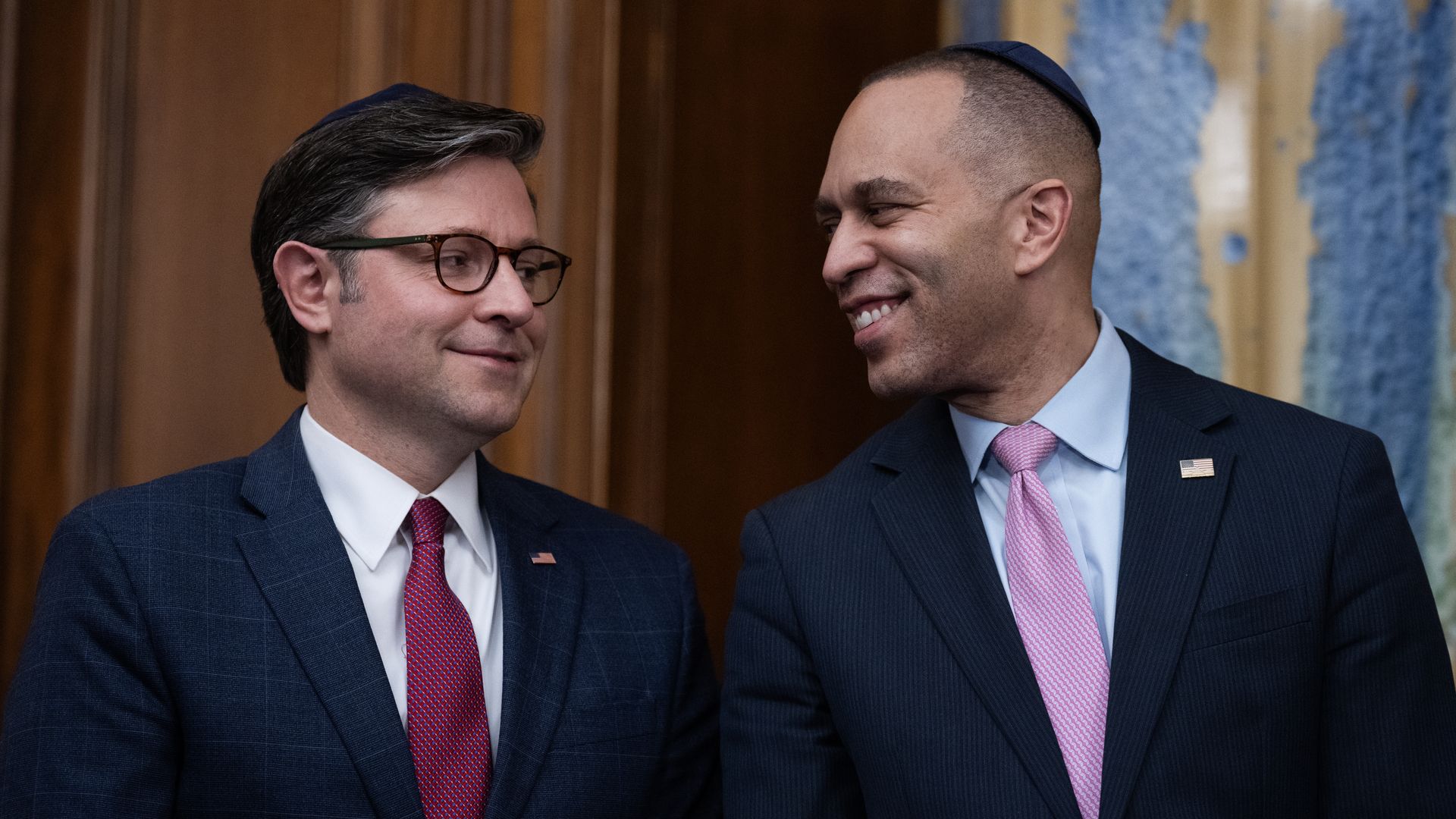Two men wearing suits and ties, one in a red tie and glasses, the other in a pink tie and a kippah, smiling at each other indoors.