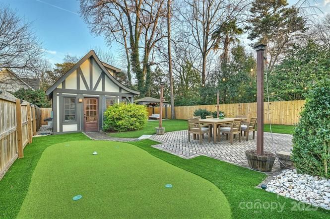 Backyard with small Tudor-style house, putting green, wooden table and chairs on brick patio, tall trees, wooden fence, and green bushes under clear sky.