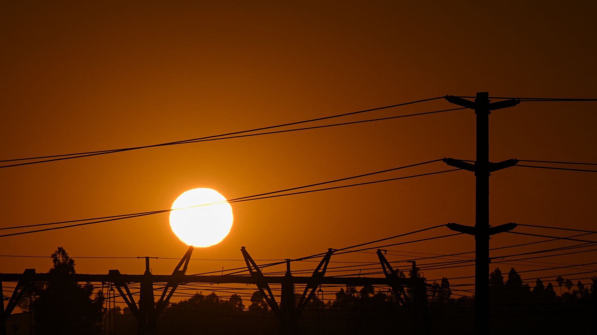 Silhouetted utility pole with multiple power lines across a warm orange sunset; dark fence silhouettes and distant trees in the foreground.
