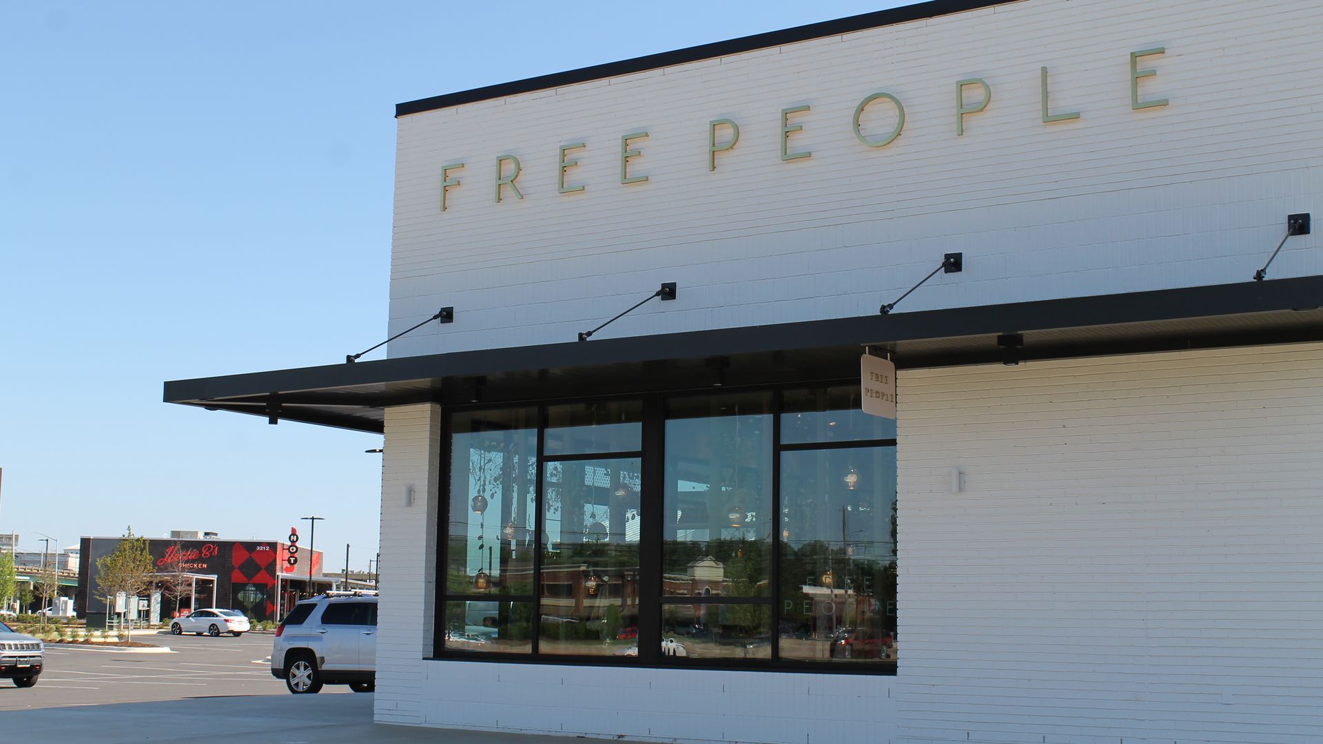White brick storefront with large glass windows and a black overhang; gold letters spell "FREE PEOPLE" on the facade. Blue sky, a parking lot, and distant buildings in the background.