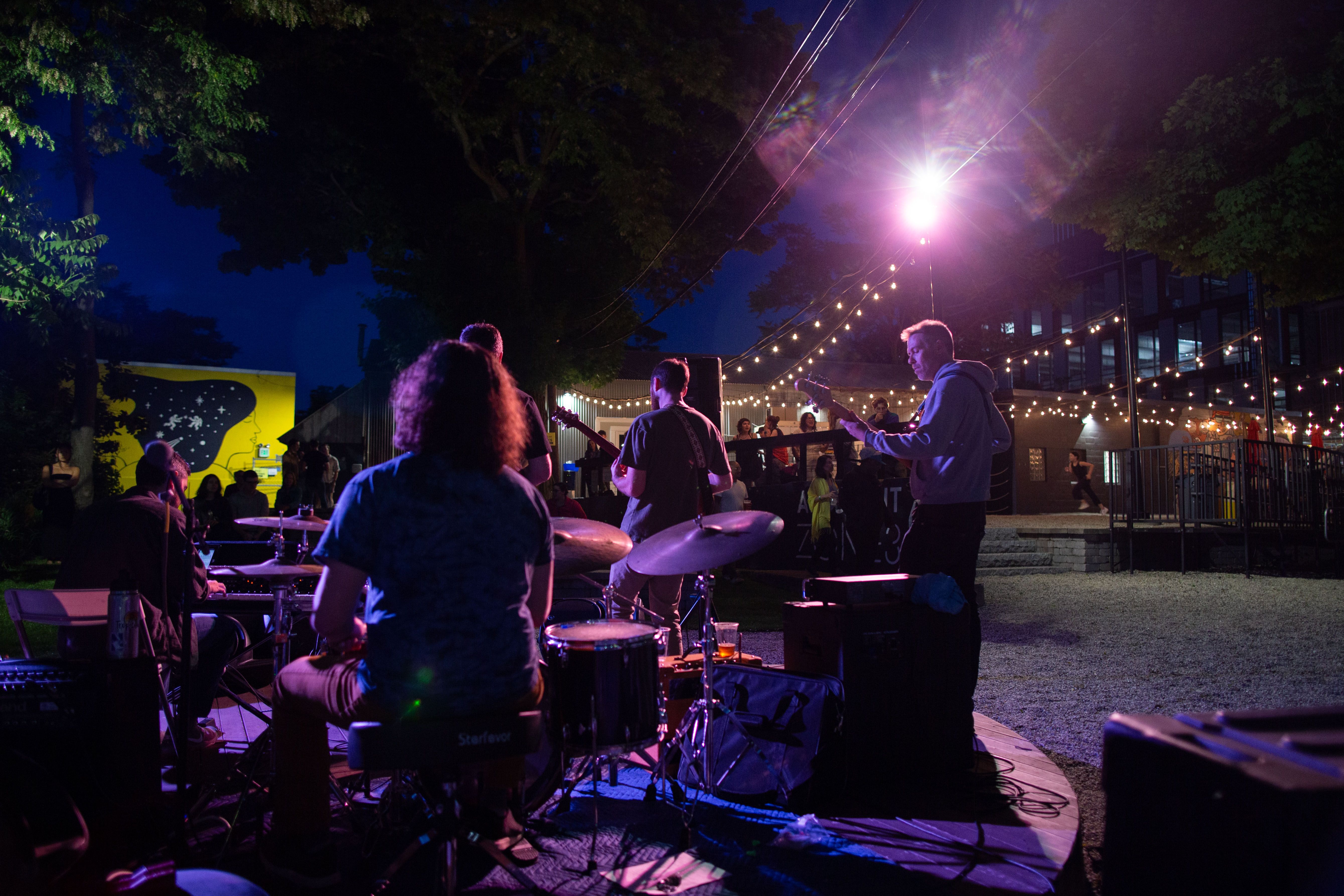 A live band, with a drummer on a drum set and two guitarists, perform outside at Aeronaut Allston one night. 
