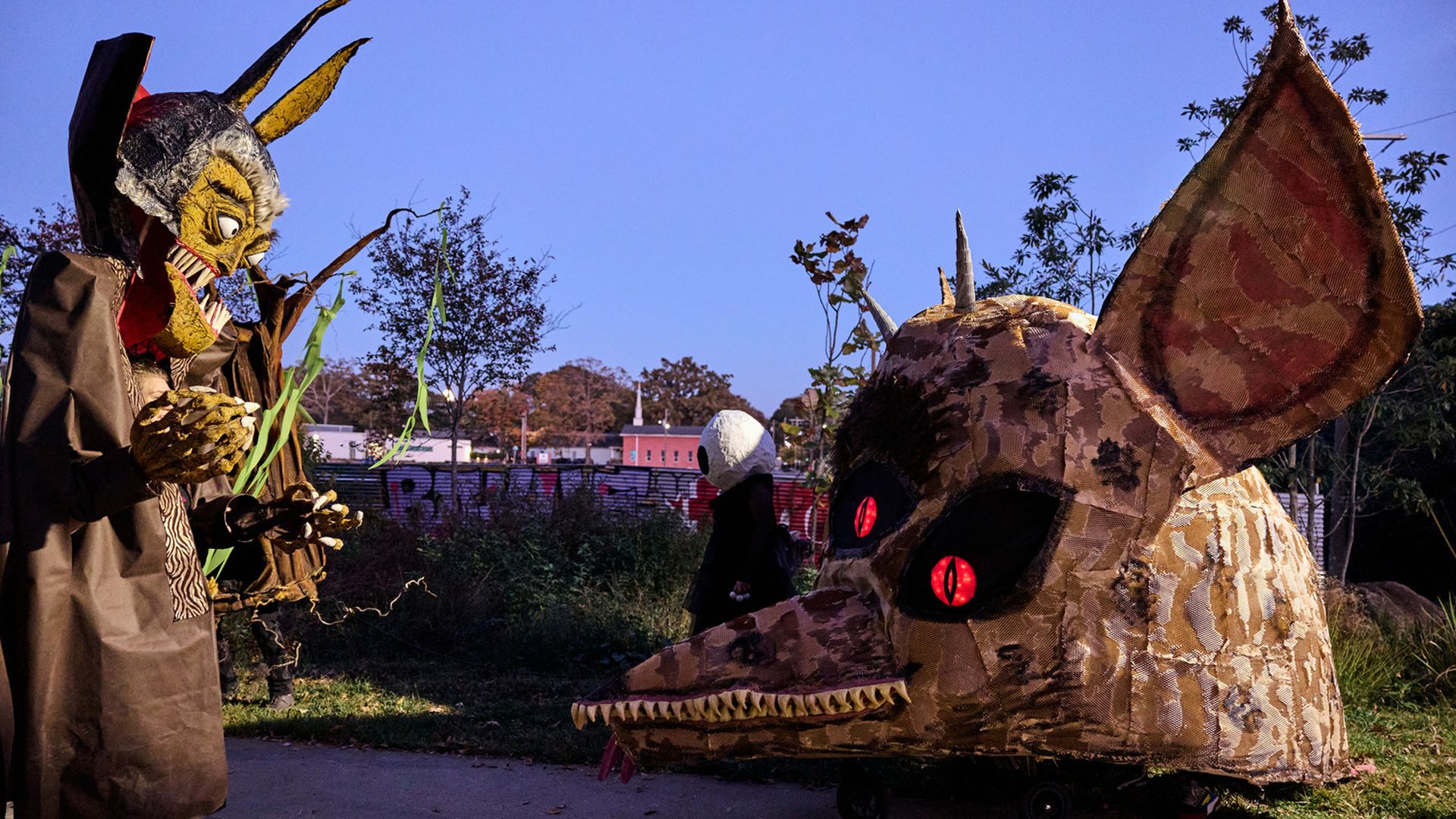 Two people in large, detailed creature costumes outdoors at twilight. One wears a tall yellow and brown demon mask; the other is a large brown creature head with red eyes and big ears.