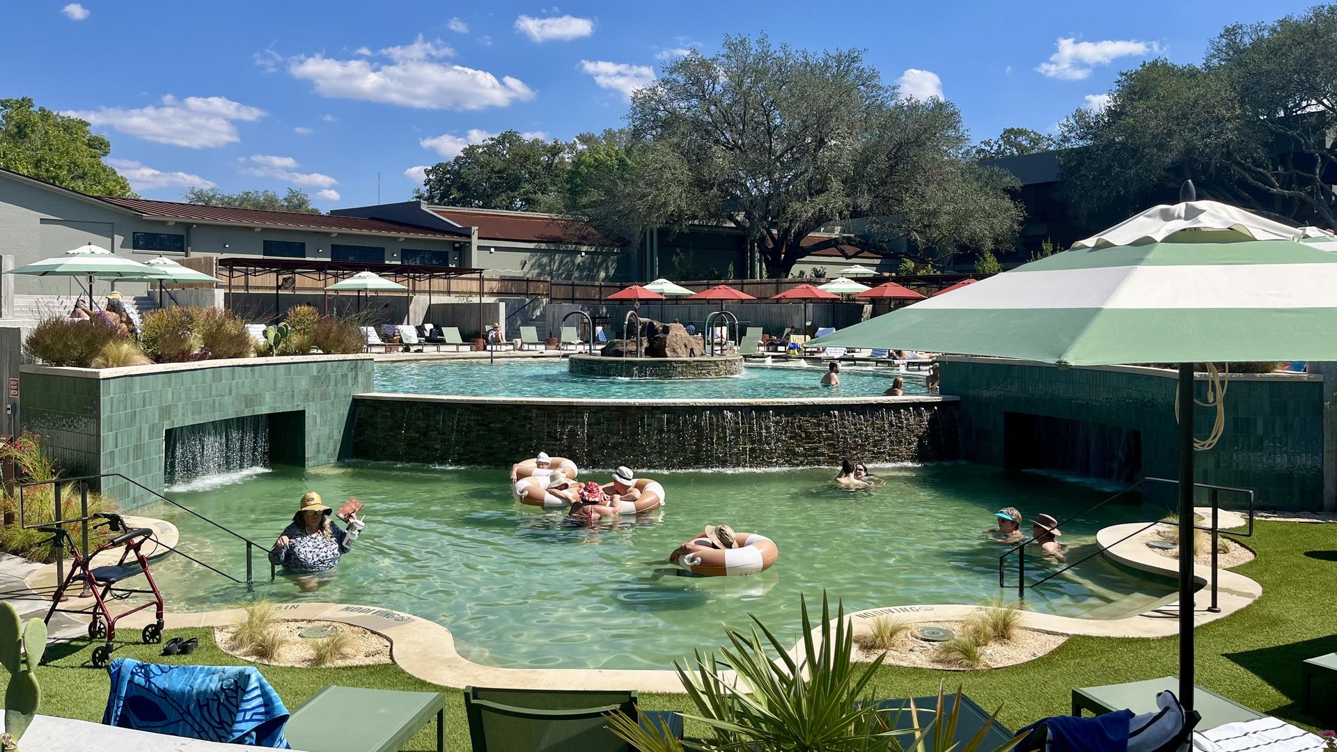 People relaxing in a greenish-blue pool with waterfalls, surrounded by green and white umbrellas, lawn chairs, and greenery under a bright blue sky with scattered white clouds.