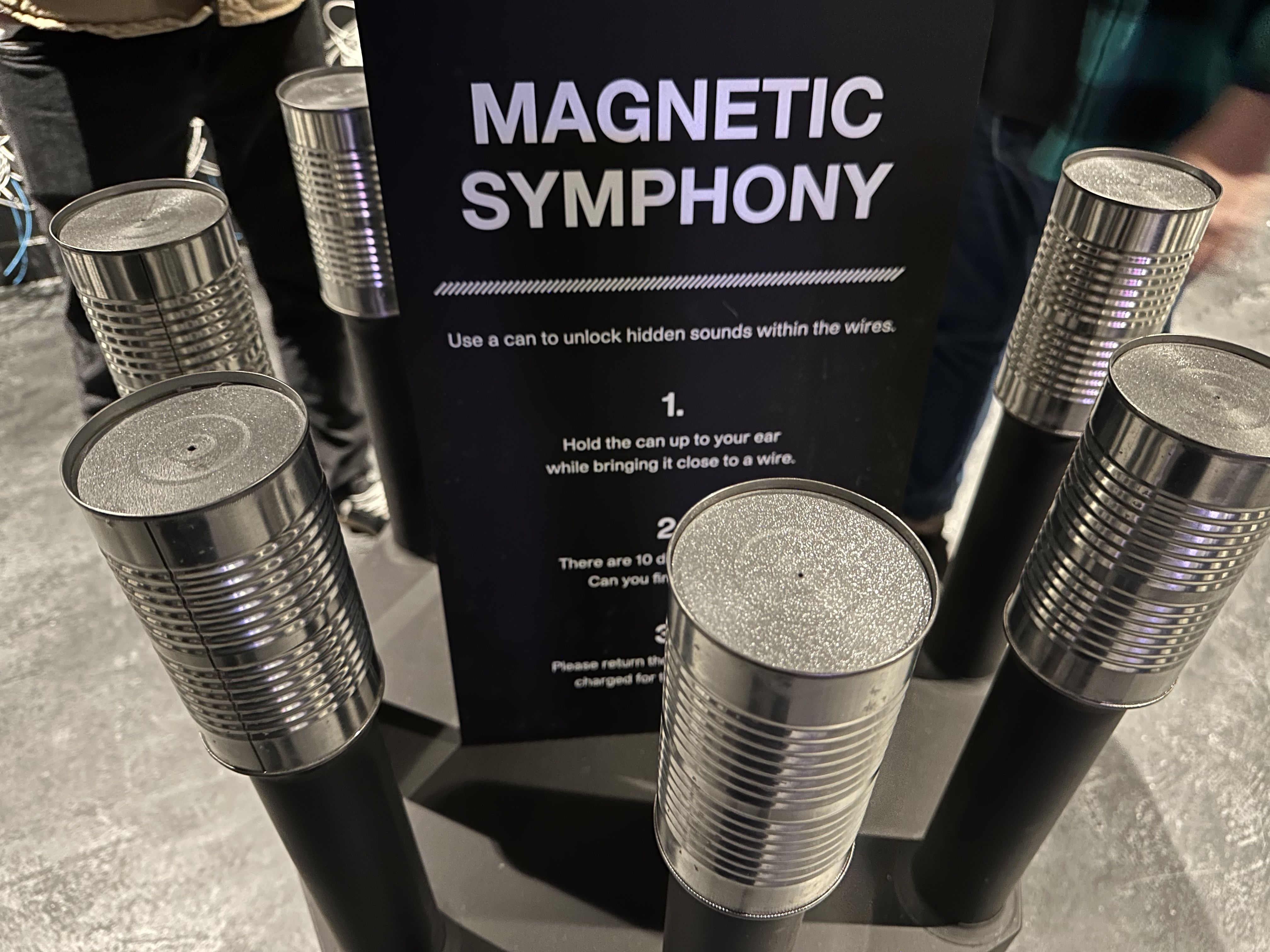 Several tin cans with electronic devices sit in a circle around a sign that shows how to use the electronic cans (you hold them up to a wire, and it plays a sound).