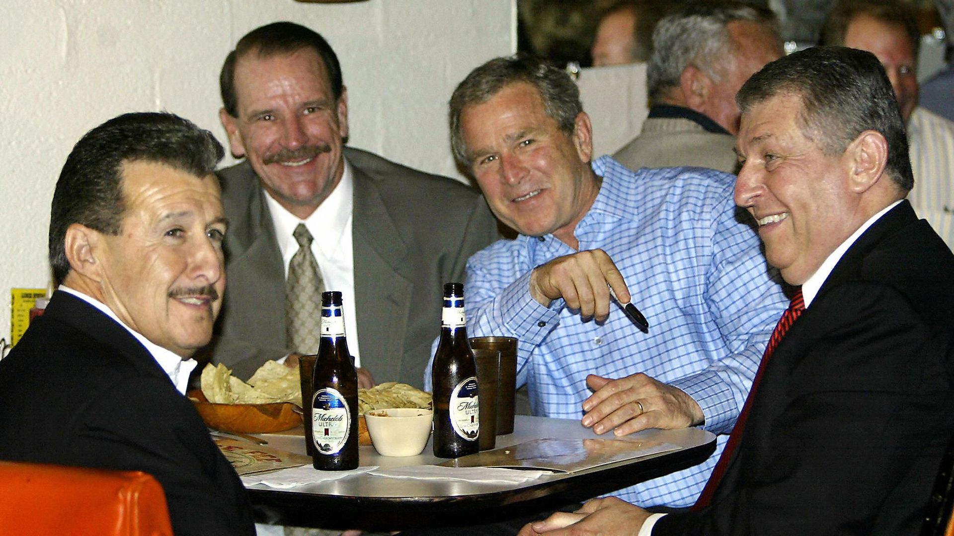 Four men sit around a table with chips and two beer bottles, smiling; a man in a blue check shirt points with a pen while the others look on.