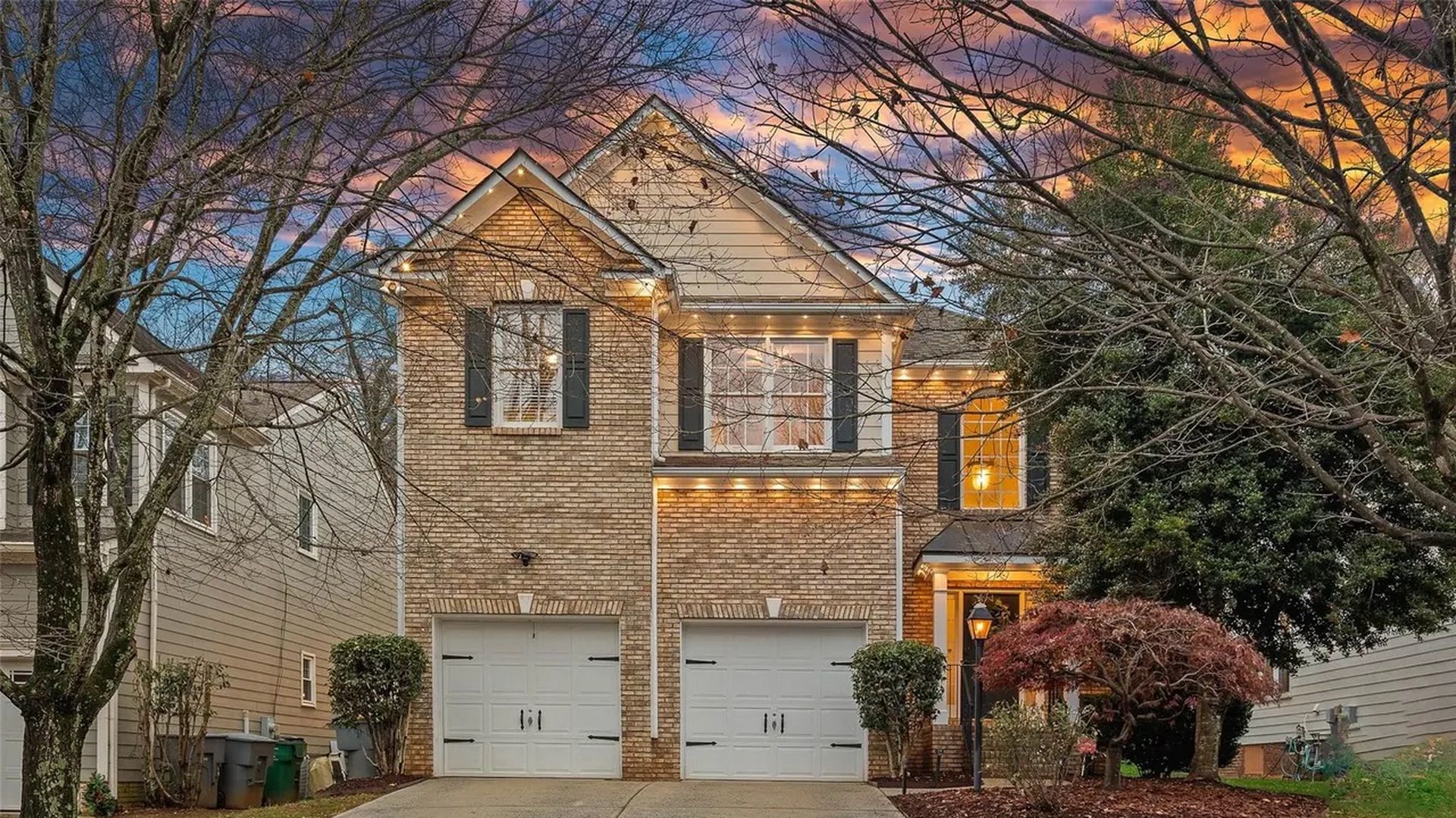 Two-story brick house with double white garage doors, lit warmly at dusk, leafless trees framing a colorful sunset sky, and a red bush near the entrance.