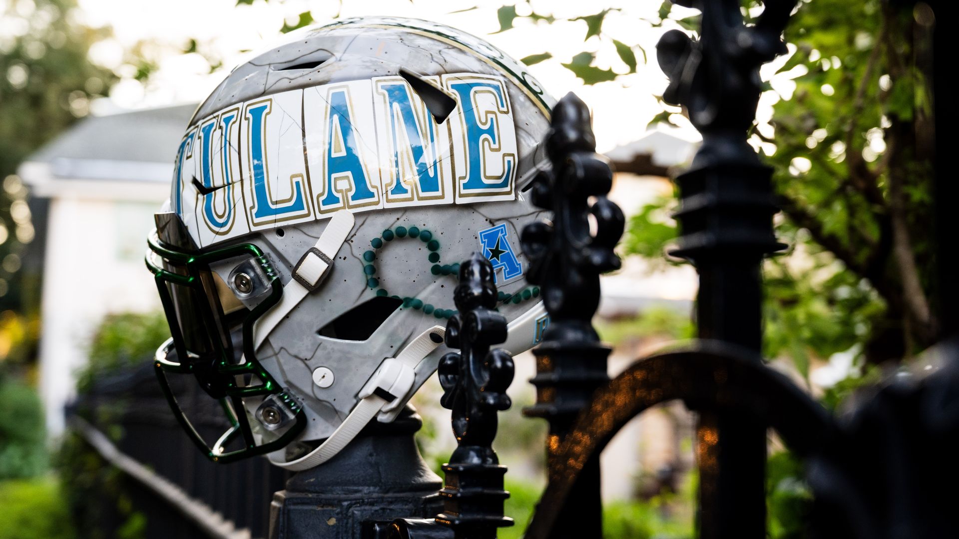 Gray football helmet with blue and gold "TULANE" letters and logo, placed on a black metal fence post, with greenery and a blurry house in the background.