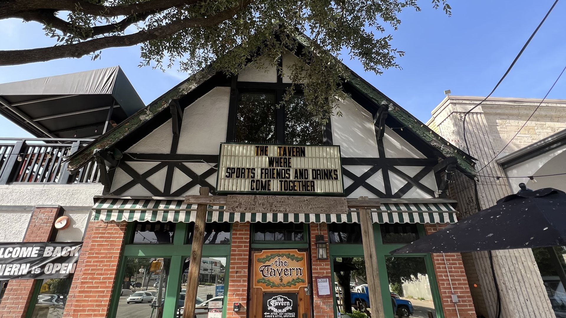 Brick building with Tudor black-and-white trim and green-framed windows sits under a large leafy tree. A striped green awning covers the storefront; a marquee reads, "THE TAVERN WHERE FRIENDS AND DRINKS COME TOGETHER" against a blue sky.