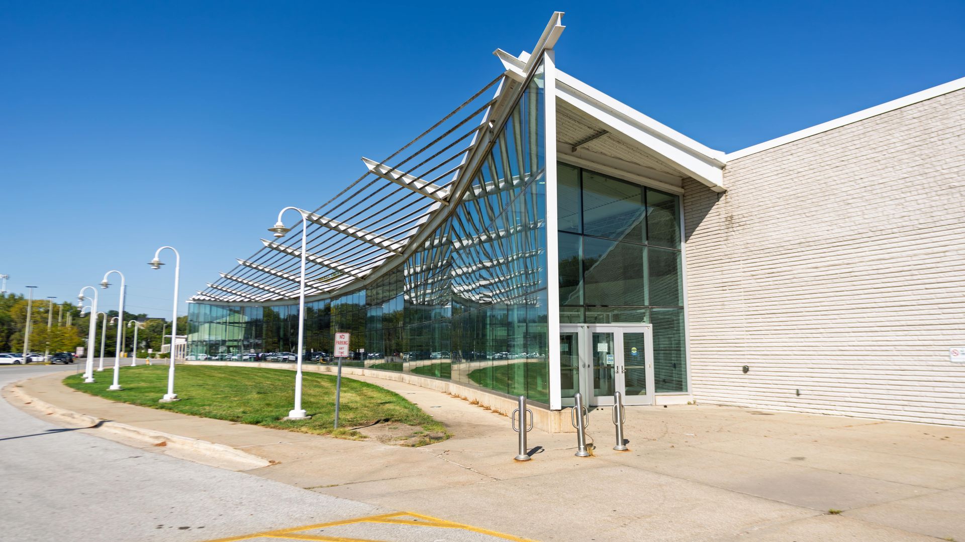 Modern building with curved glass facade and white structural beams, white brick wall, sidewalk, lampposts, green grass, and clear blue sky overhead.