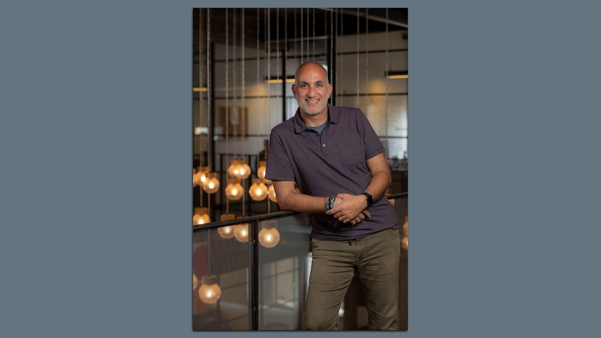 Smiling middle-aged man in a dark purple polo shirt leans on a railing inside a modern office, with hanging globe lights and glass walls in the background.