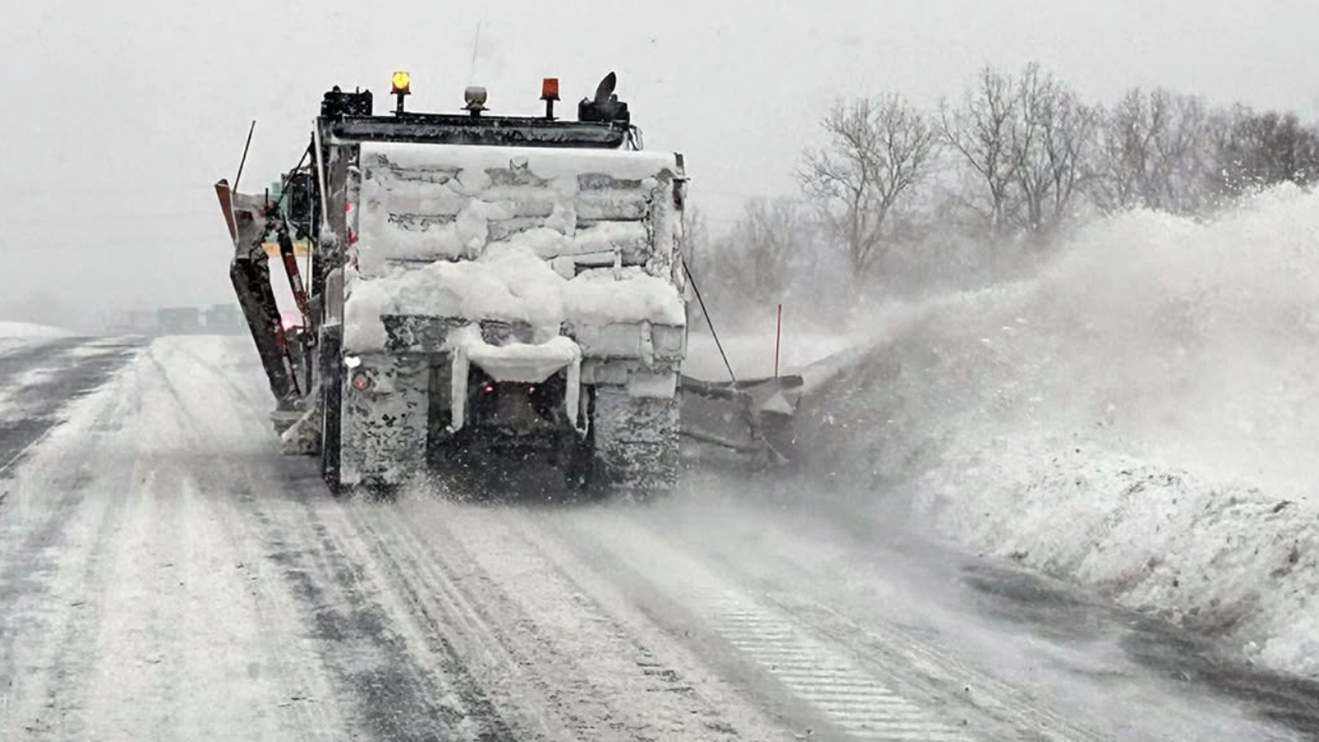 Snowplow truck clearing snow from a road during a snowy, gray winter day, with snow being pushed to the side and bare trees in the background.