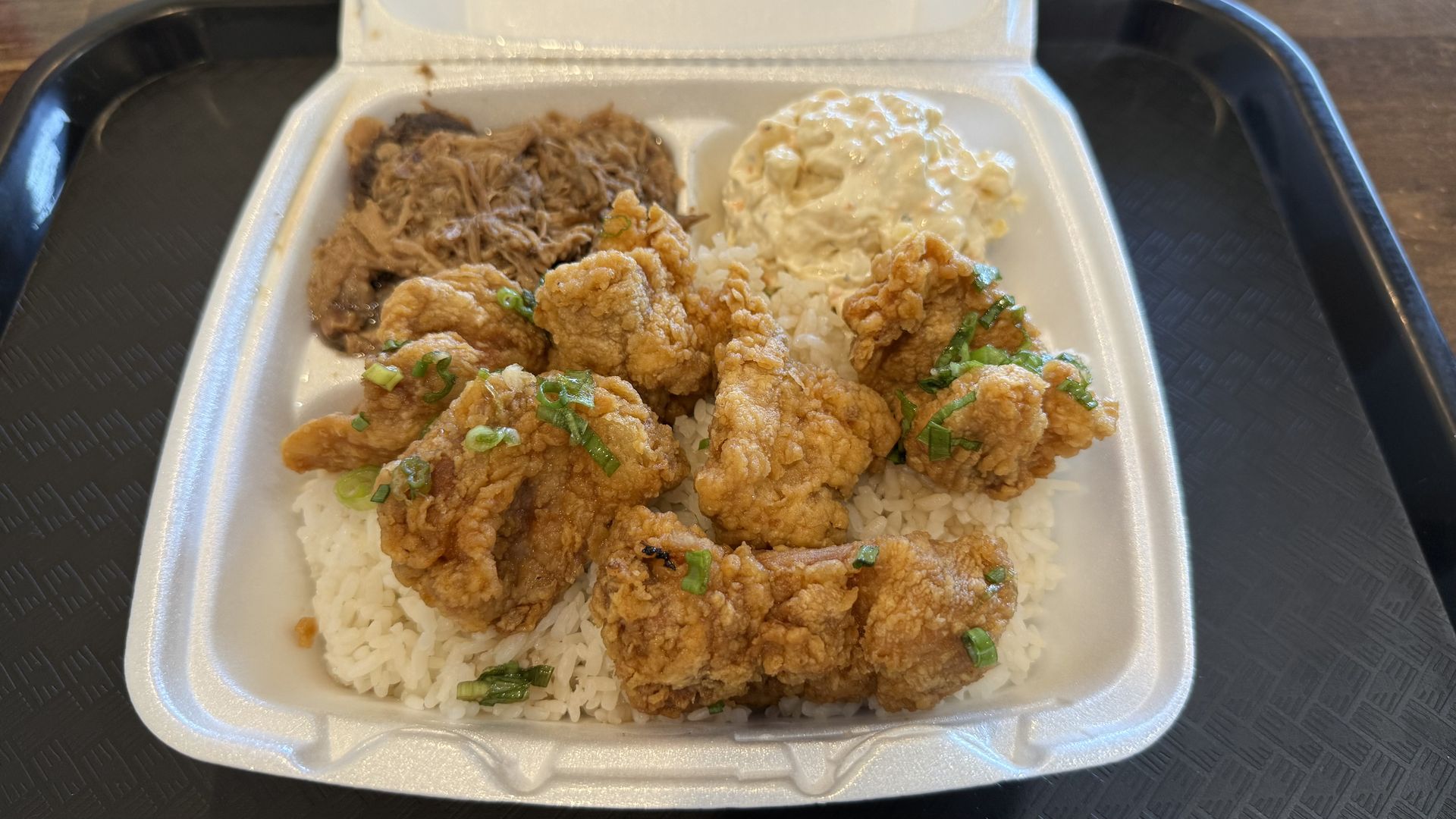 A compartmentalized takeout container with white rice topped with fried chicken pieces garnished with green onions, a scoop of macaroni salad, and shredded pork on the side.