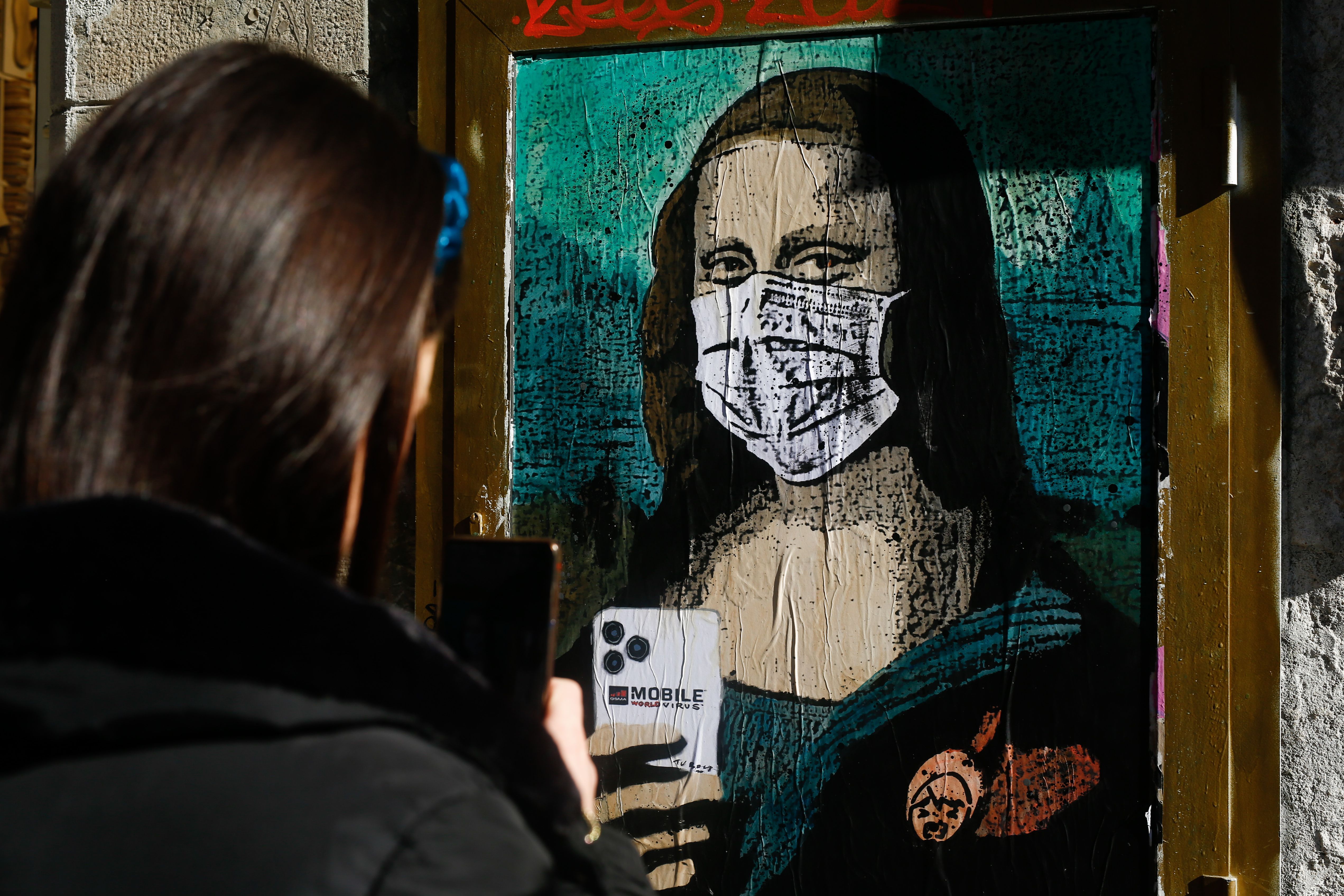 A woman takes a picture with her mobile phone of a poster by Italian urban artist Salvatore Benintende aka "TVBOY" depecting Leonardo da Vinci's Mona Lisa wearing a protective facemask 