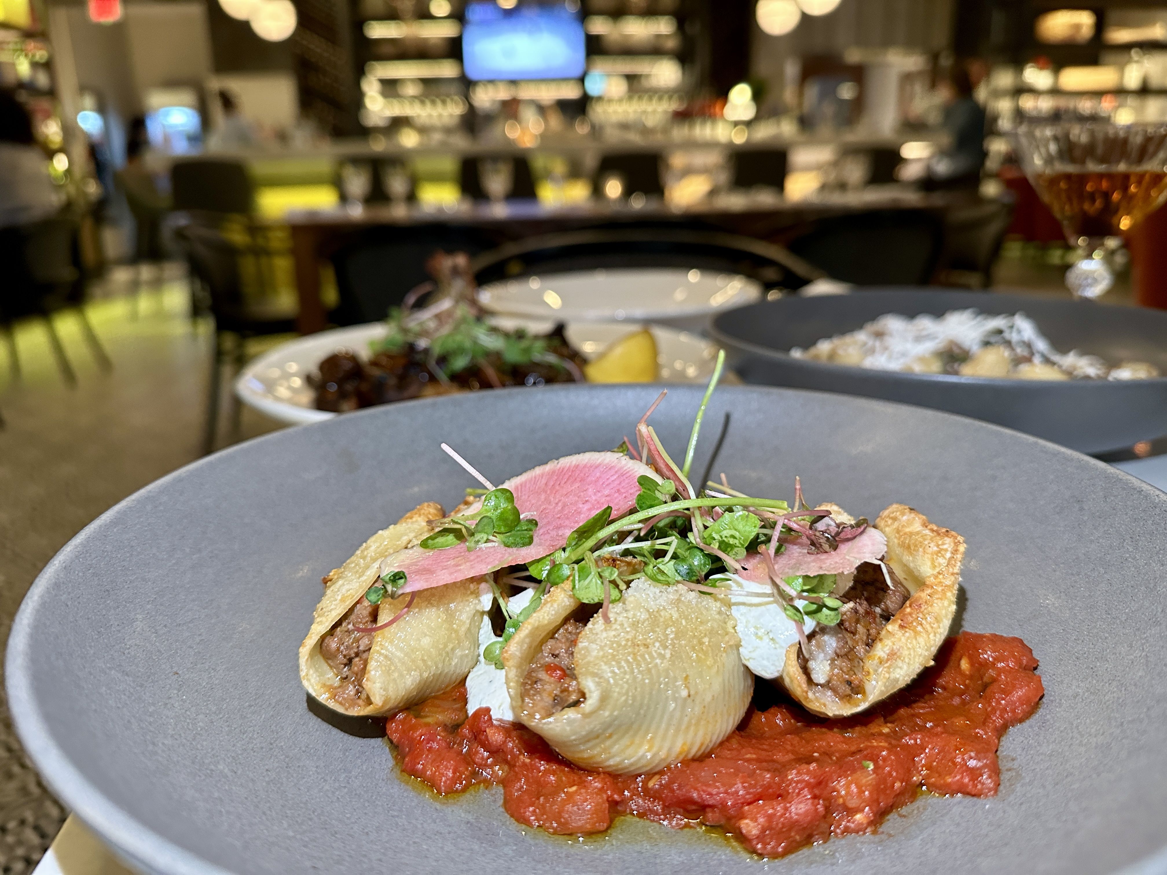 italo uptown charlotte Foreground shows a plated pasta dish with stuffed shells over red tomato sauce, topped with microgreens and a lemon wedge. Blurry restaurant bar and diners fill the background.