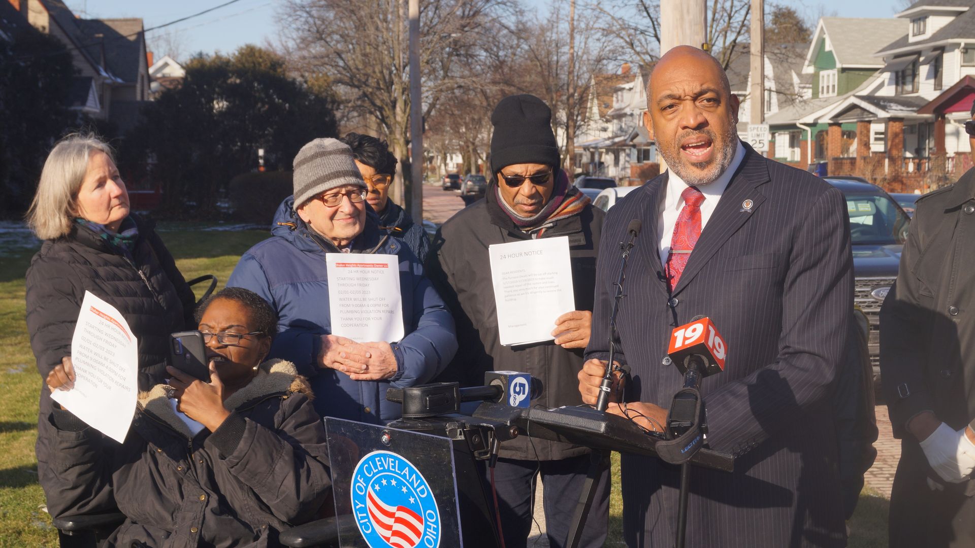 Council president Blaine Griffin speaks animatedly at a lectern outdoors, as a diverse group of neighbors in winter clothes watch