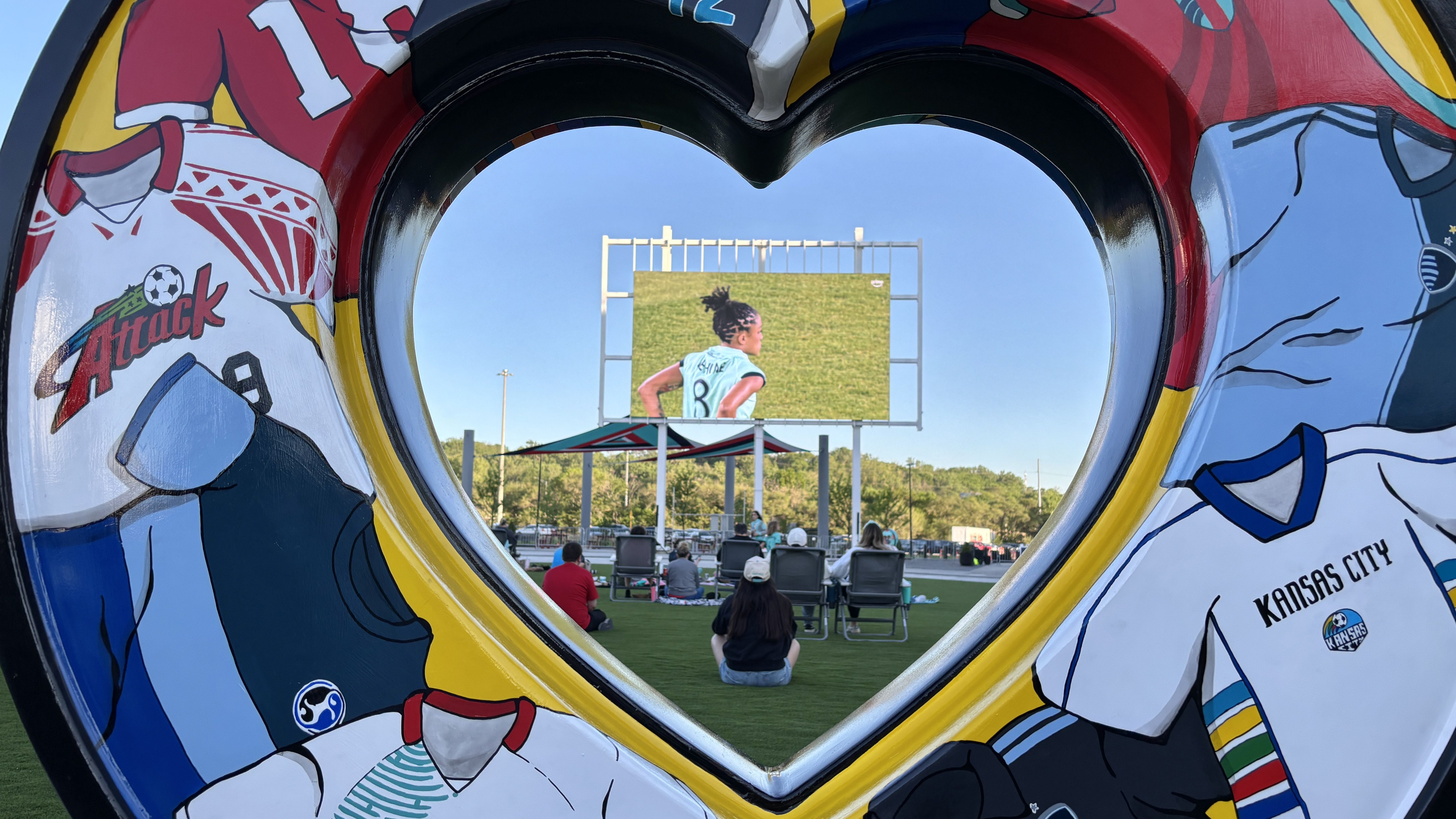 A view through a heart-shaped opening in a bright mural frame of an outdoor soccer venue: a large screen shows a female player in a light turquoise jersey #8 on a green field under a blue sky; spectators sit nearby.