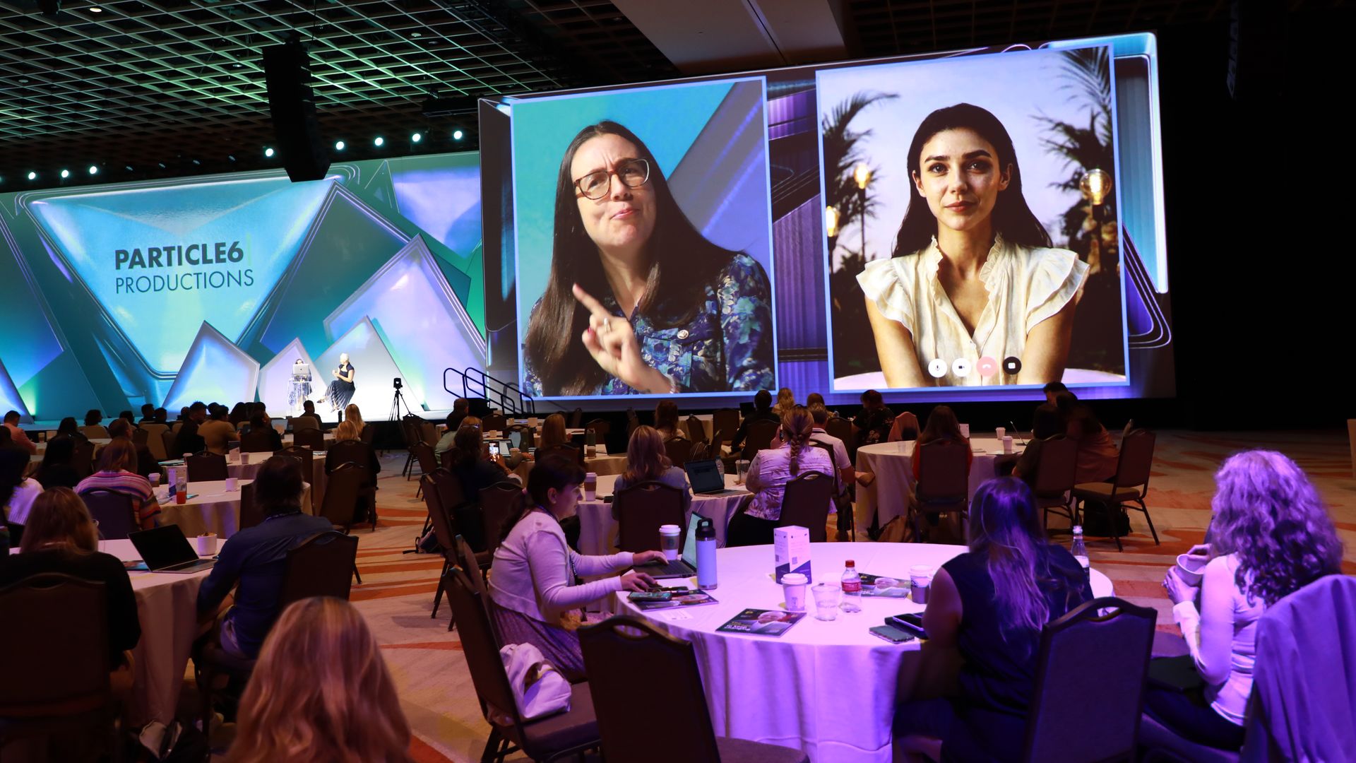 Conference hall stage with large screens showing two women; left panel has a woman with glasses, right panel shows a woman outdoors; audience sits at round tables; "Particle6 Productions" logo on the left.