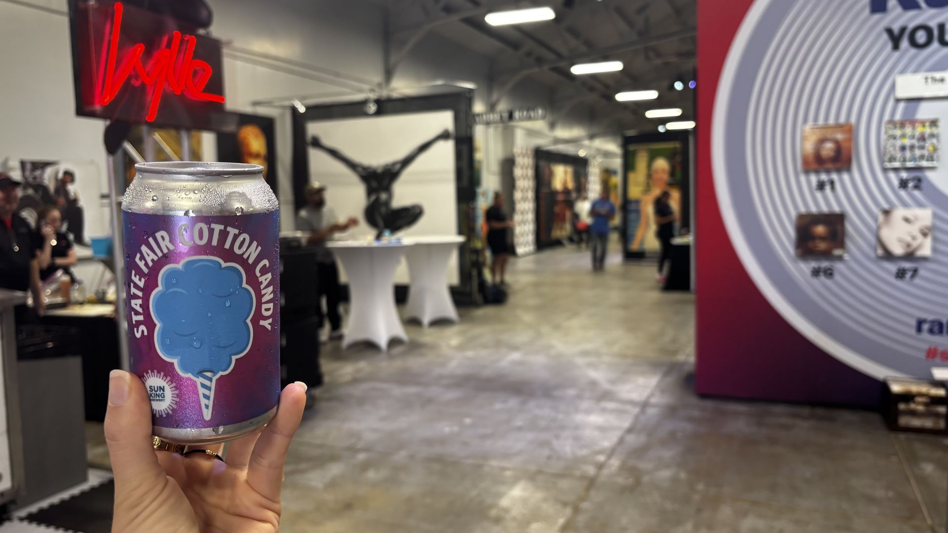 Hand holding a wet can labeled "State Fair Cotton Candy" with a blue cotton candy image at an indoor event with people, artwork, and booths in the background.