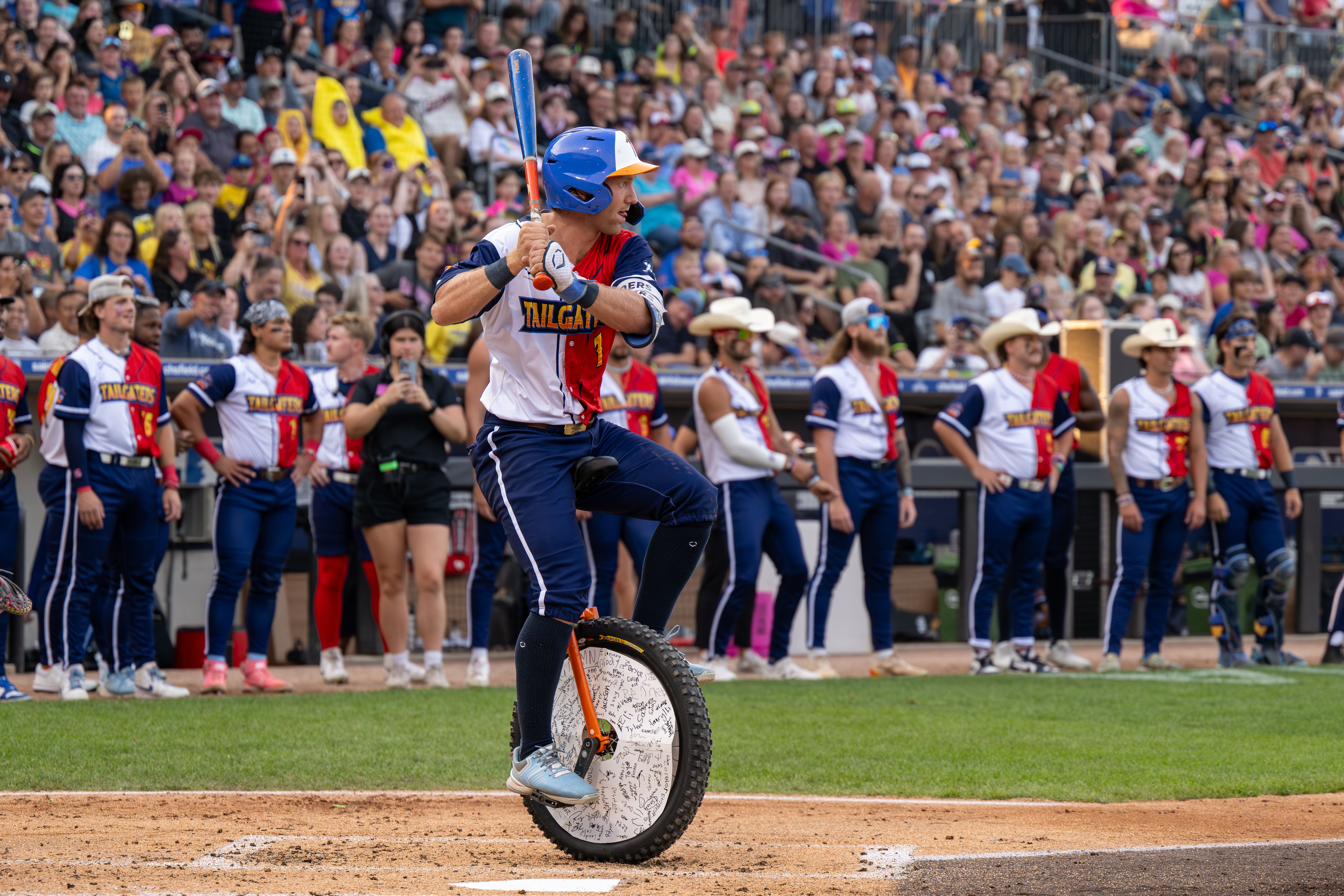 Baseball player in red, white, and blue uniform rides a unicycle at home plate, holding a bat; teammates and crowd watch in a lively stadium setting.