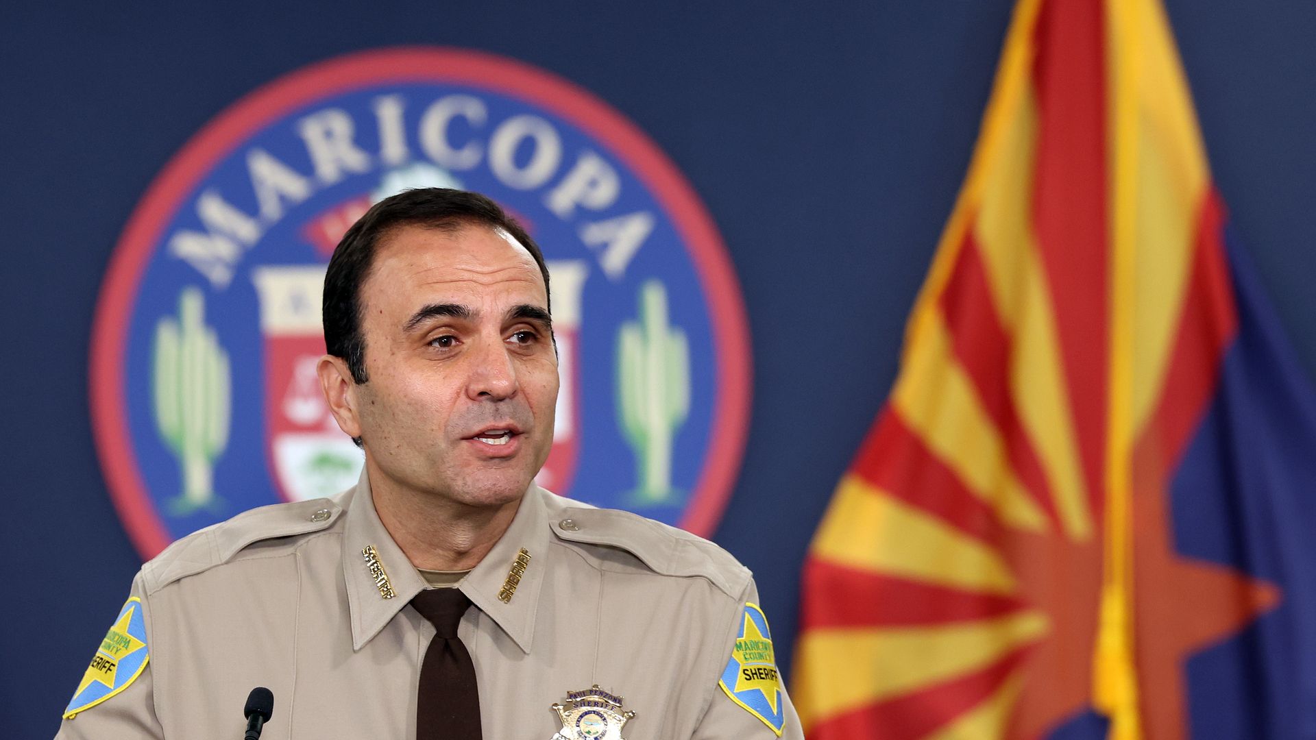 A man in a beige law enforcement uniform speaks in front of a large emblem that says Maricopa.
