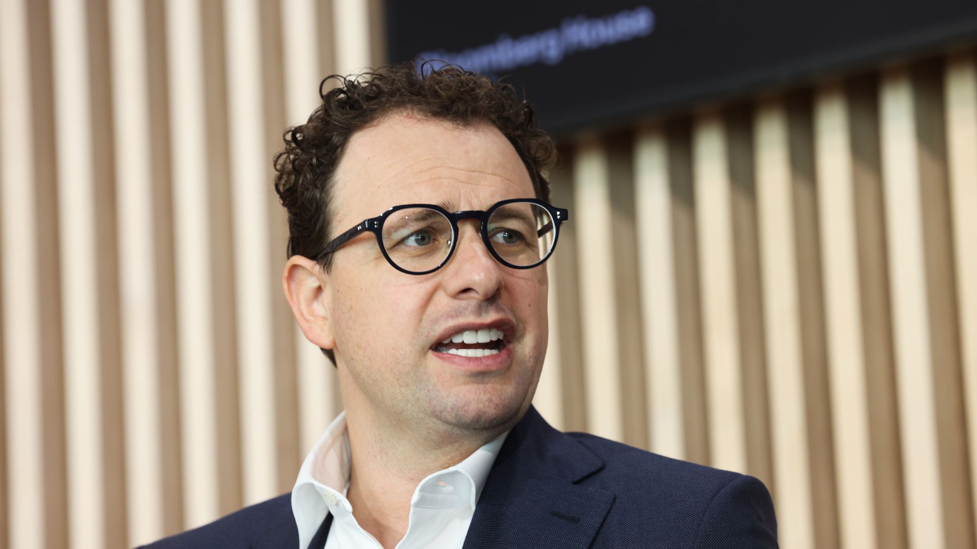 A man with curly hair and round black glasses wearing a navy blazer and white shirt, speaking in front of vertical beige blinds.