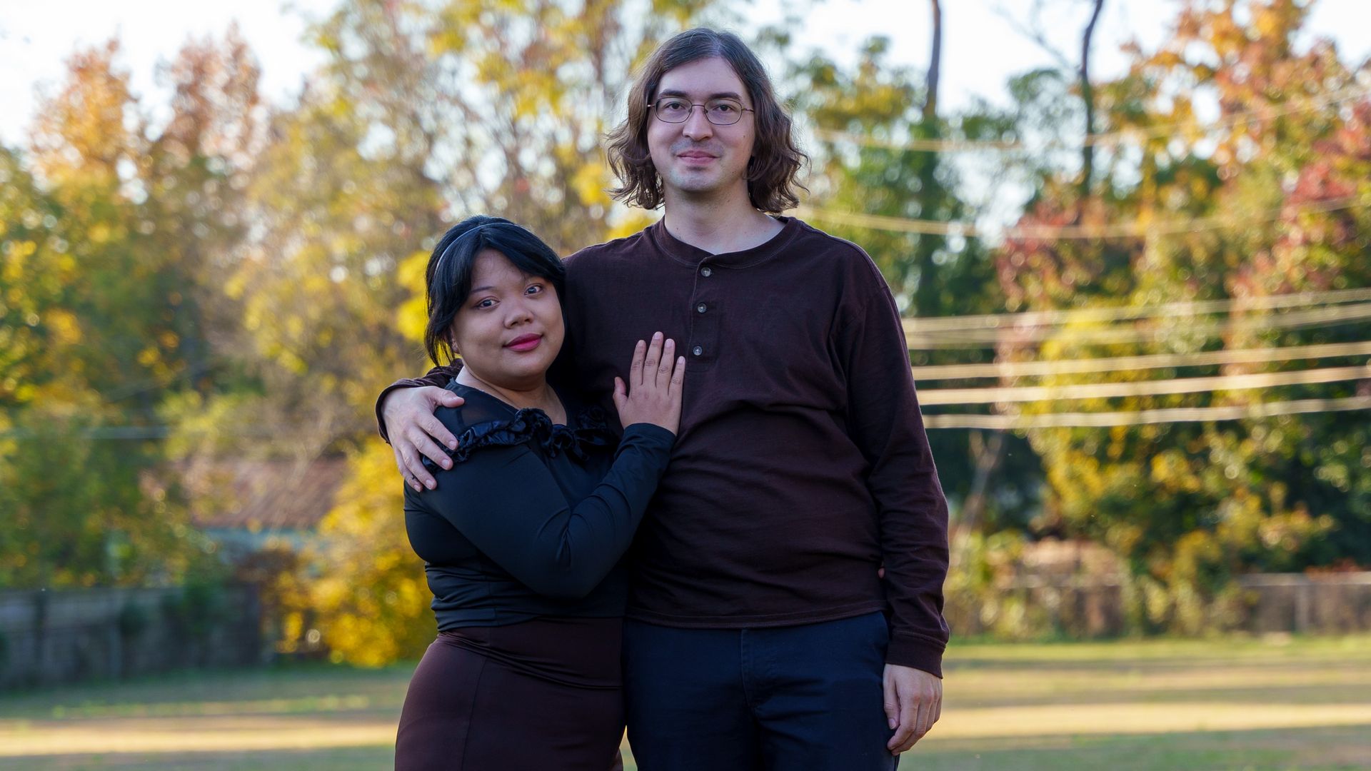 Two people stand arm in arm in a park on a sunny autumn day. The woman wears a dark outfit and leans into the taller man in a brown shirt and dark pants, with colorful fall trees behind them.