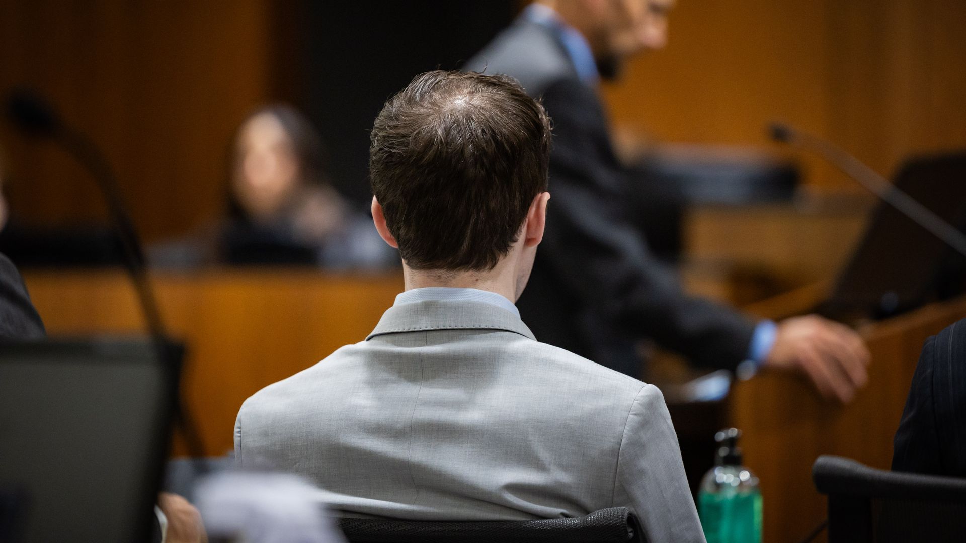 Back of a person with short brown hair in a light gray suit, seated in a courtroom; a blurred man at a podium in the background, wooden walls, and a green water bottle in the foreground.