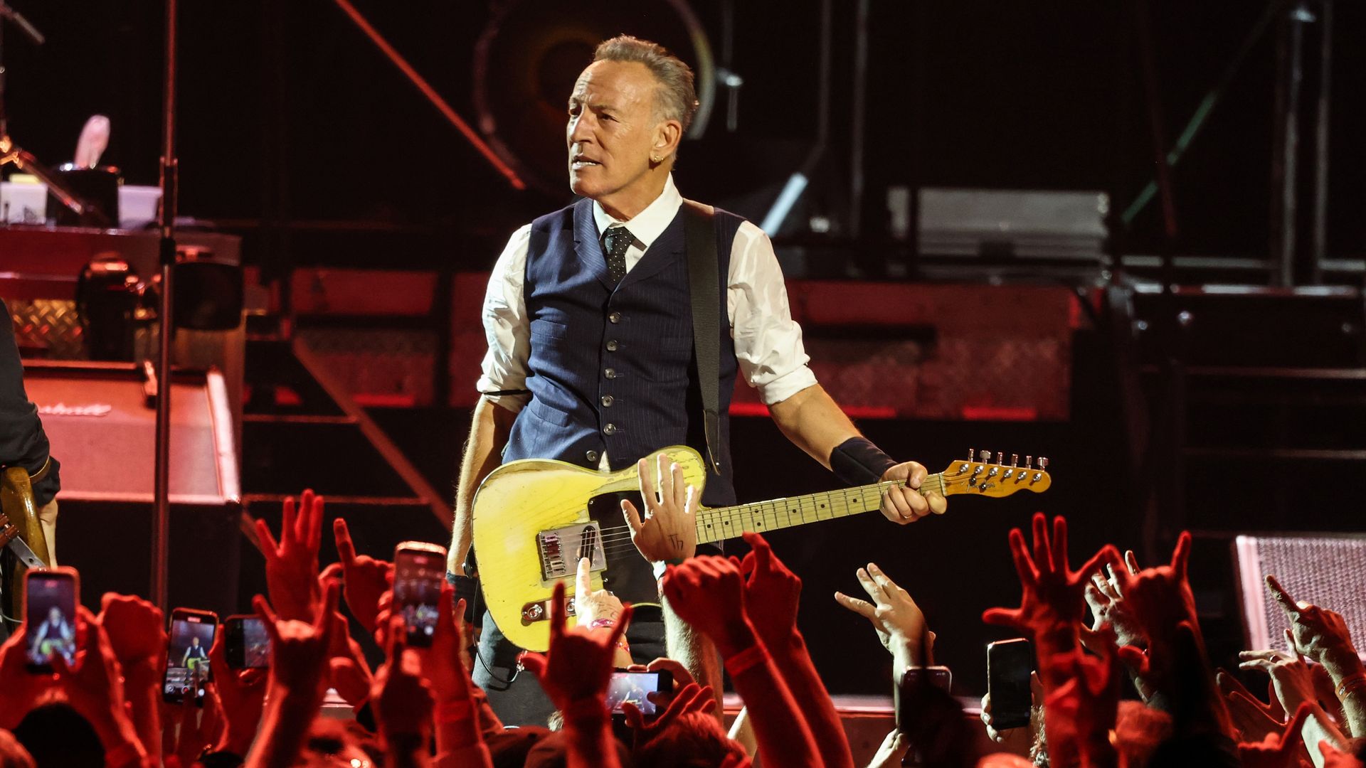 Male guitarist in a navy blue vest and white shirt plays a yellow electric guitar on a dim stage, as a cheering crowd raises hands and smartphones in the foreground.