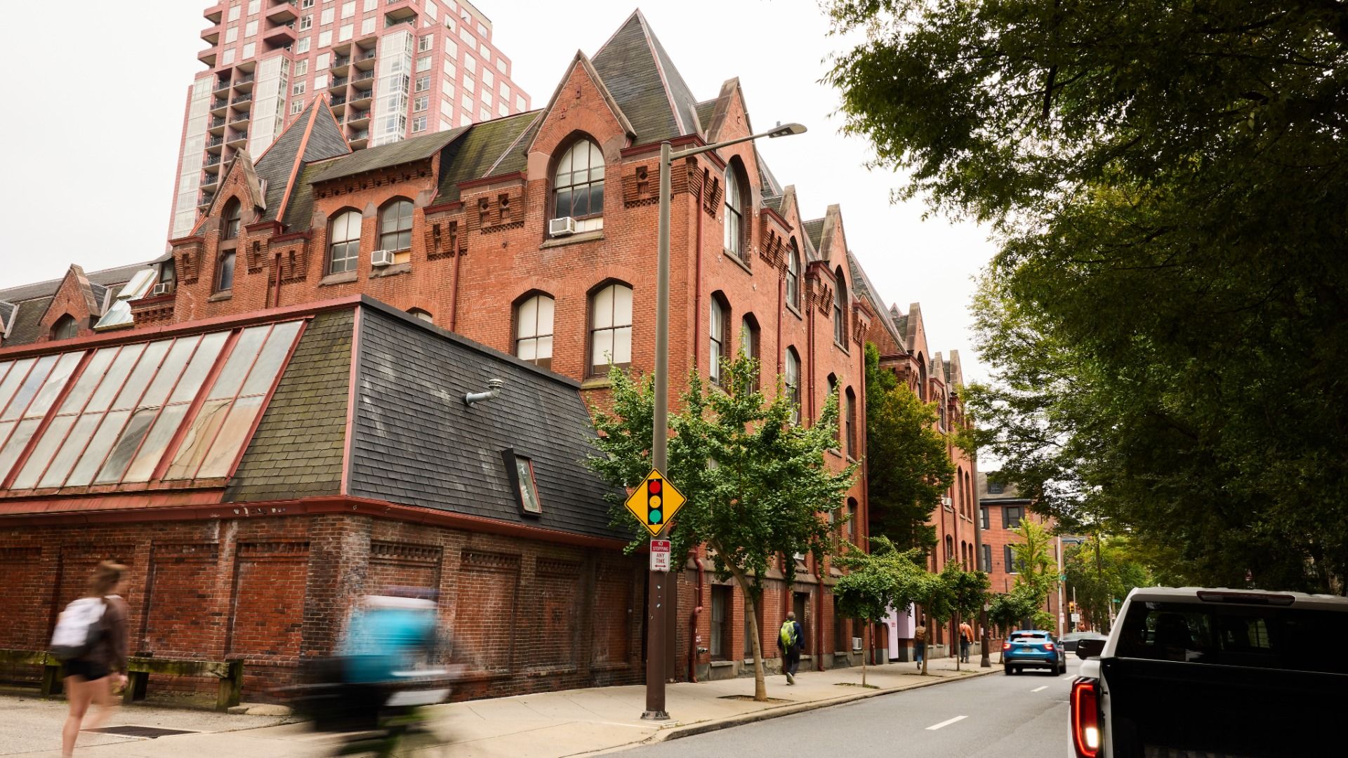 Red brick building with arched windows on a city street, beside a tall modern high-rise. Green trees line the right; a traffic light sign and blurred pedestrians and cars move along the road.