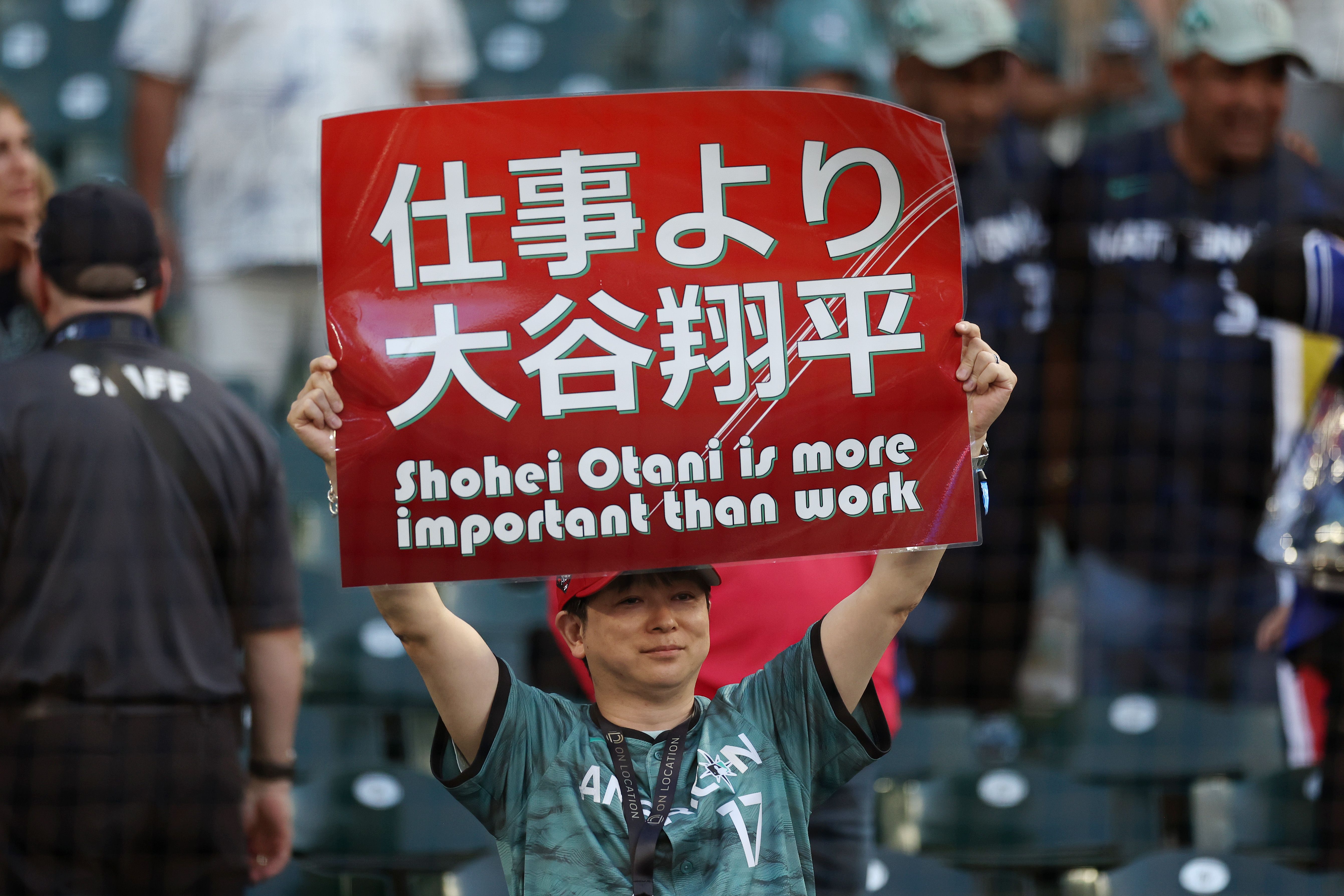 A person in a green jersey and red hat holds a sign that says "Shohei Otani is more important than work."