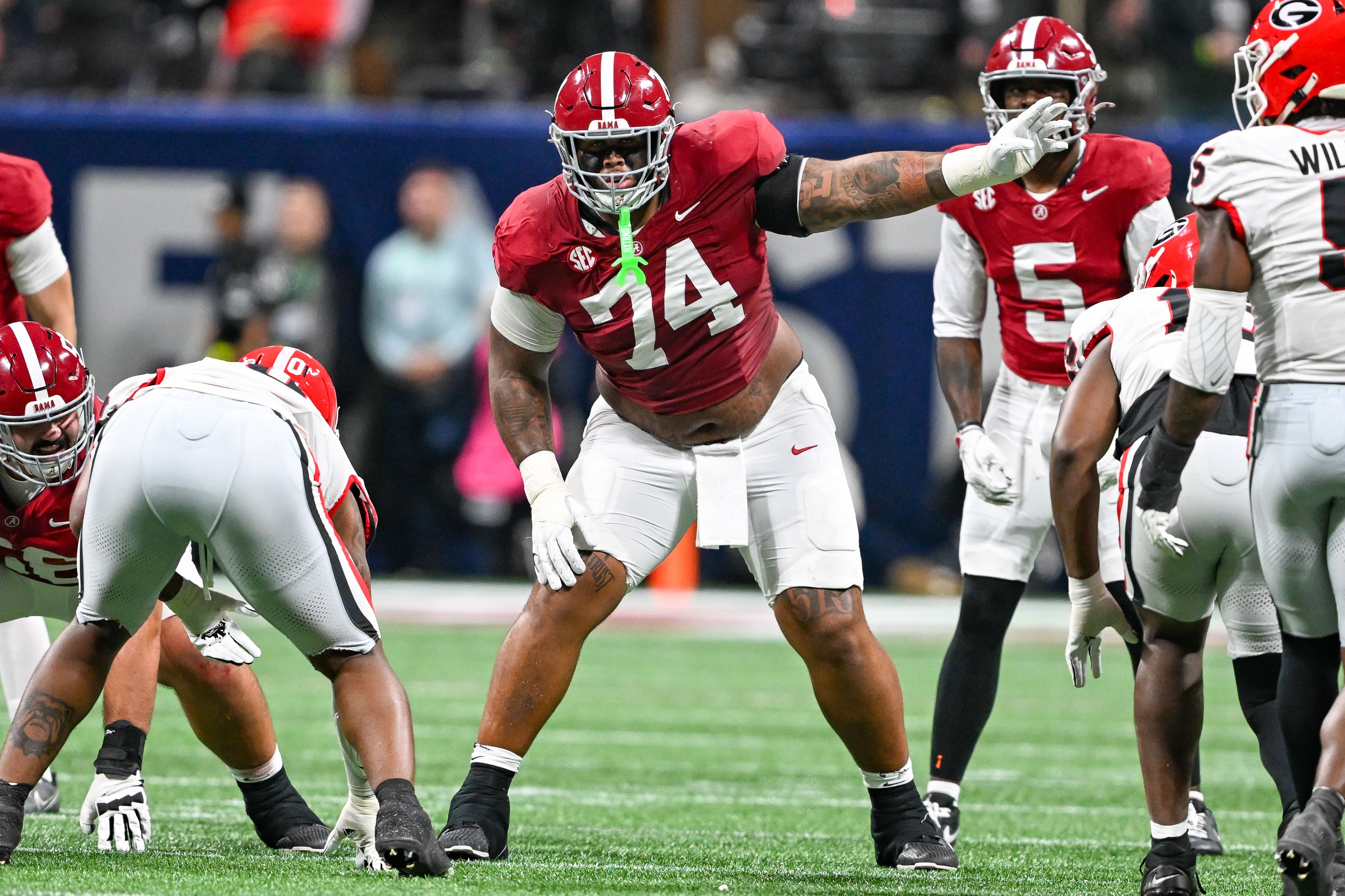 Alabama football players in red jerseys and white pants on a green field during a game; centered, #74 lunges forward with outstretched arm, neon mouthguard visible; teammates nearby.
