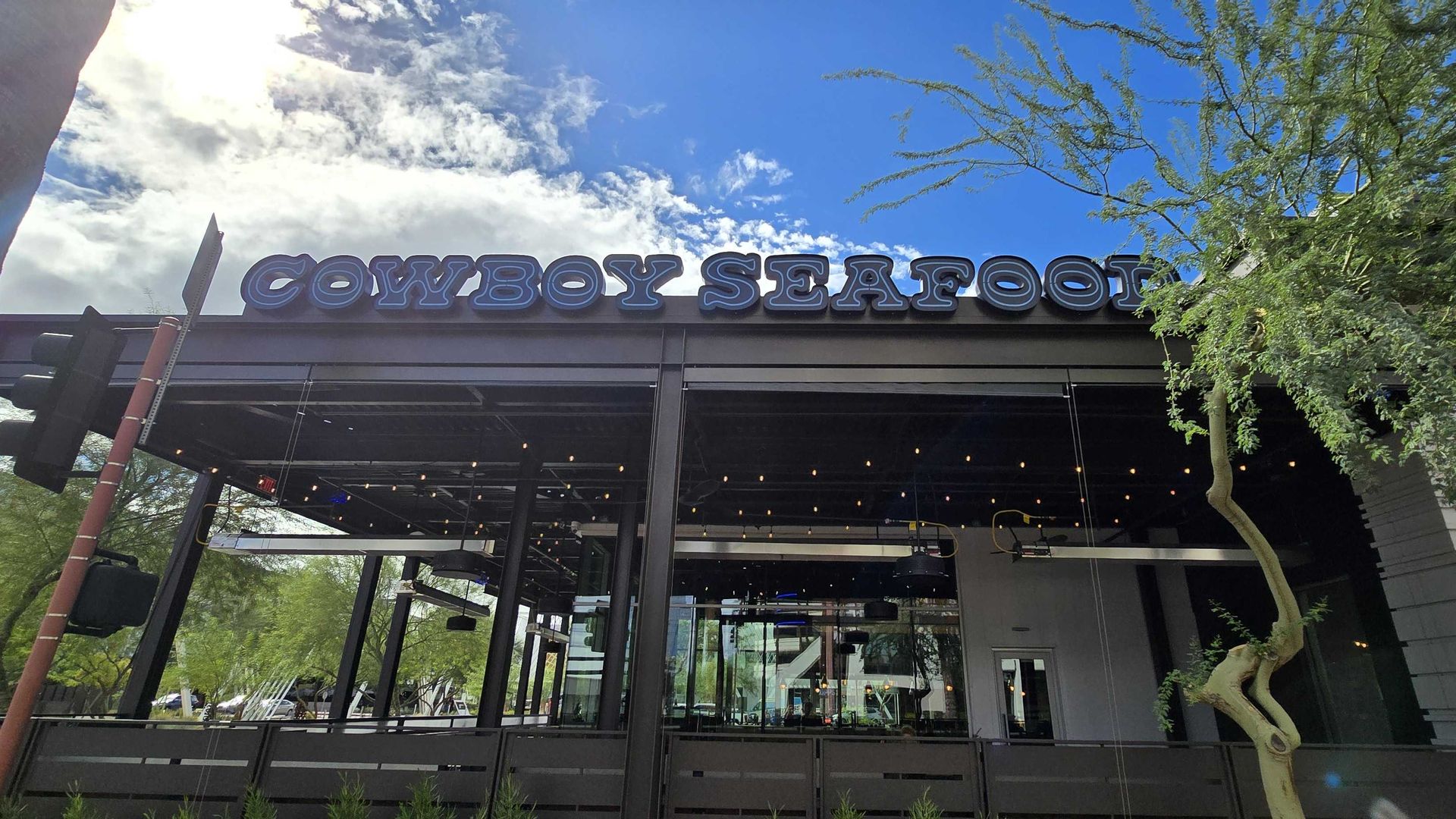 Storefront with a large blue sign reading "COWBOY SEAFOOD" above a glass entrance. Bright blue sky with clouds, sun flare on the left, and green trees on the right; dark modern facade.