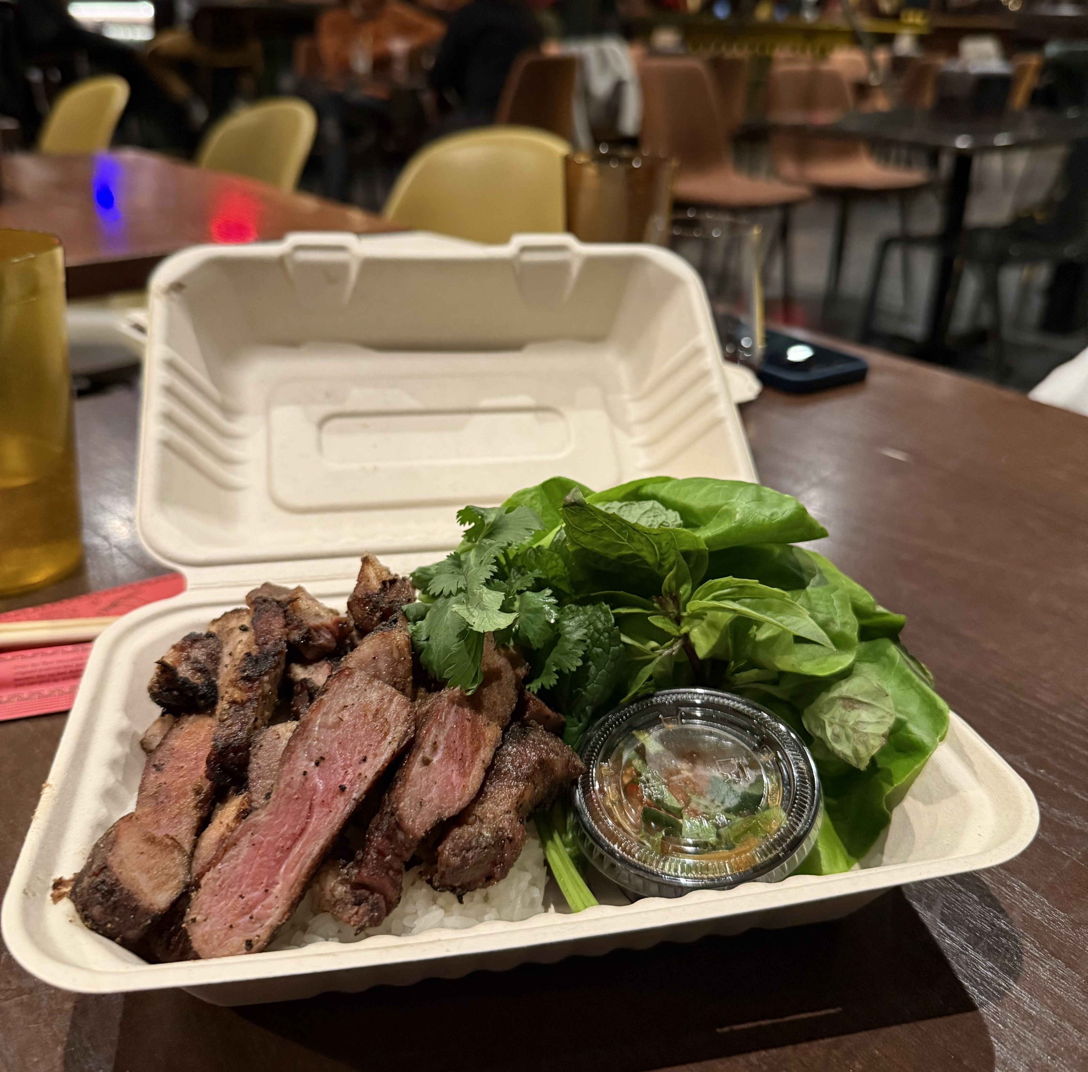 Takeout container with grilled medium-rare steak slices on rice, fresh green herbs, leafy greens, and a small cup of dipping sauce on a brown table in a restaurant setting.