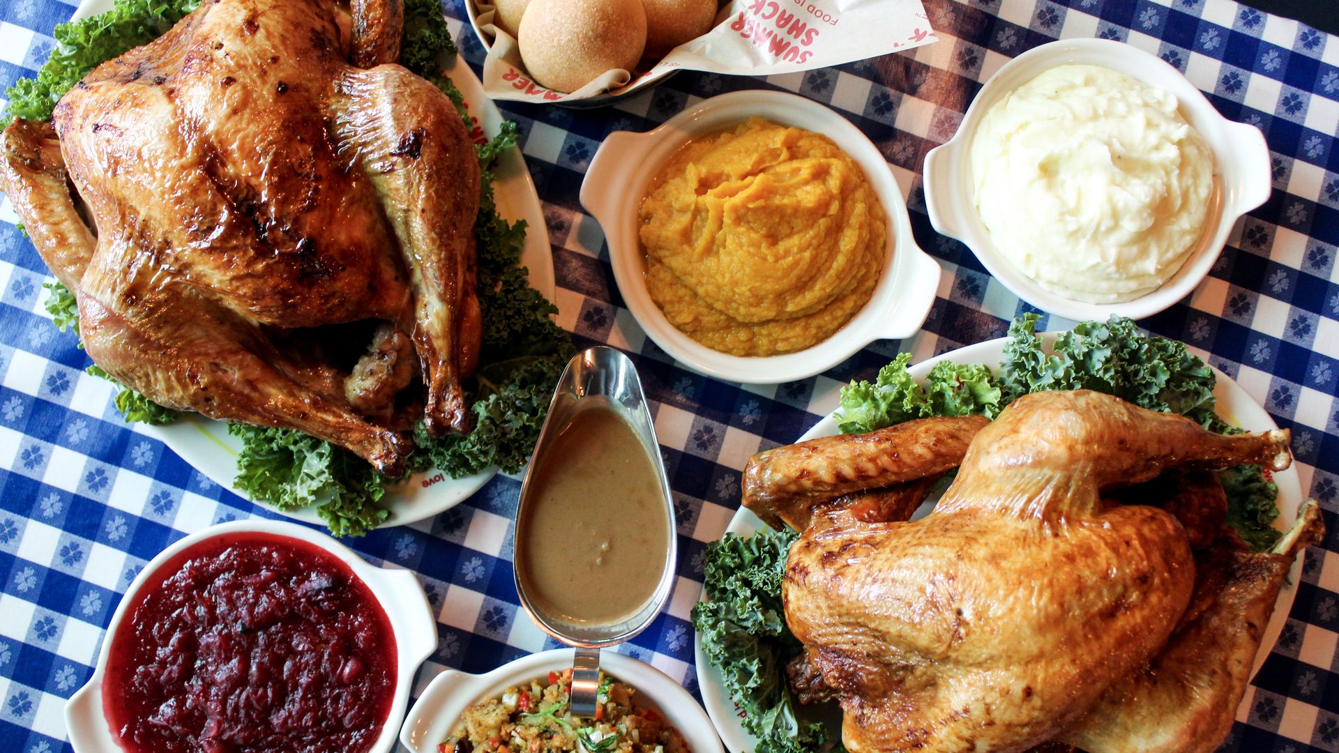 An aerial view of Thanksgiving meal on a dinner table with two roasted turkeys, mashed potatoes, gravy, sweet potatoes, bread rolls and cranberry sauce.