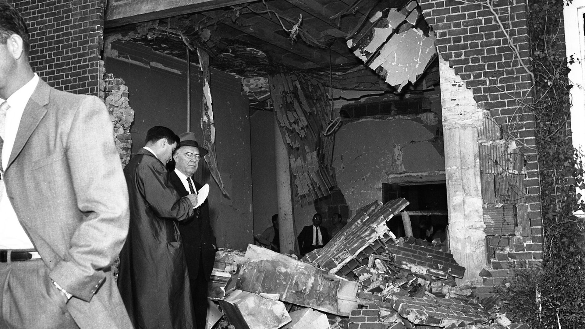 A black and white photo of two men investigating a large blast in the side of a synagogue caused by a bomb