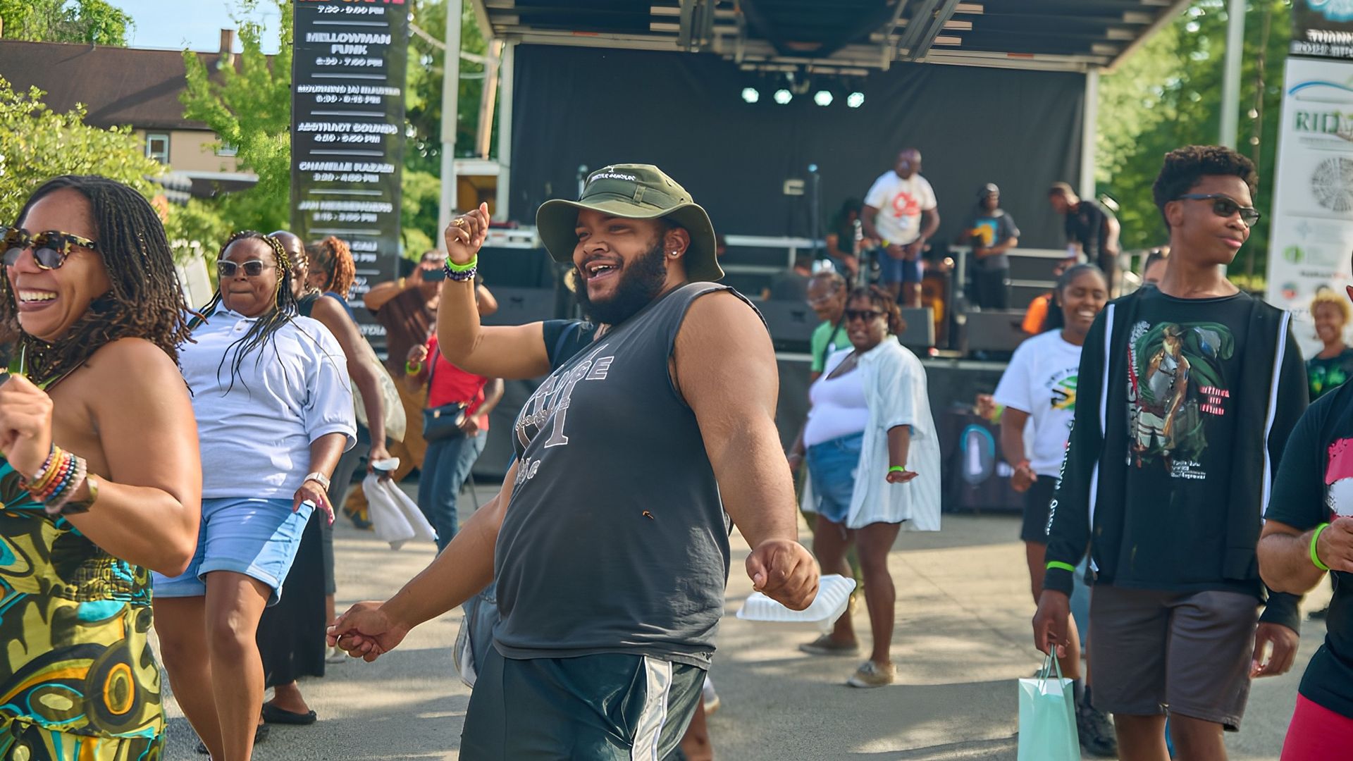 People dancing and smiling outdoors in front of a stage at a lively festival on a sunny day with green trees and a blue sky.