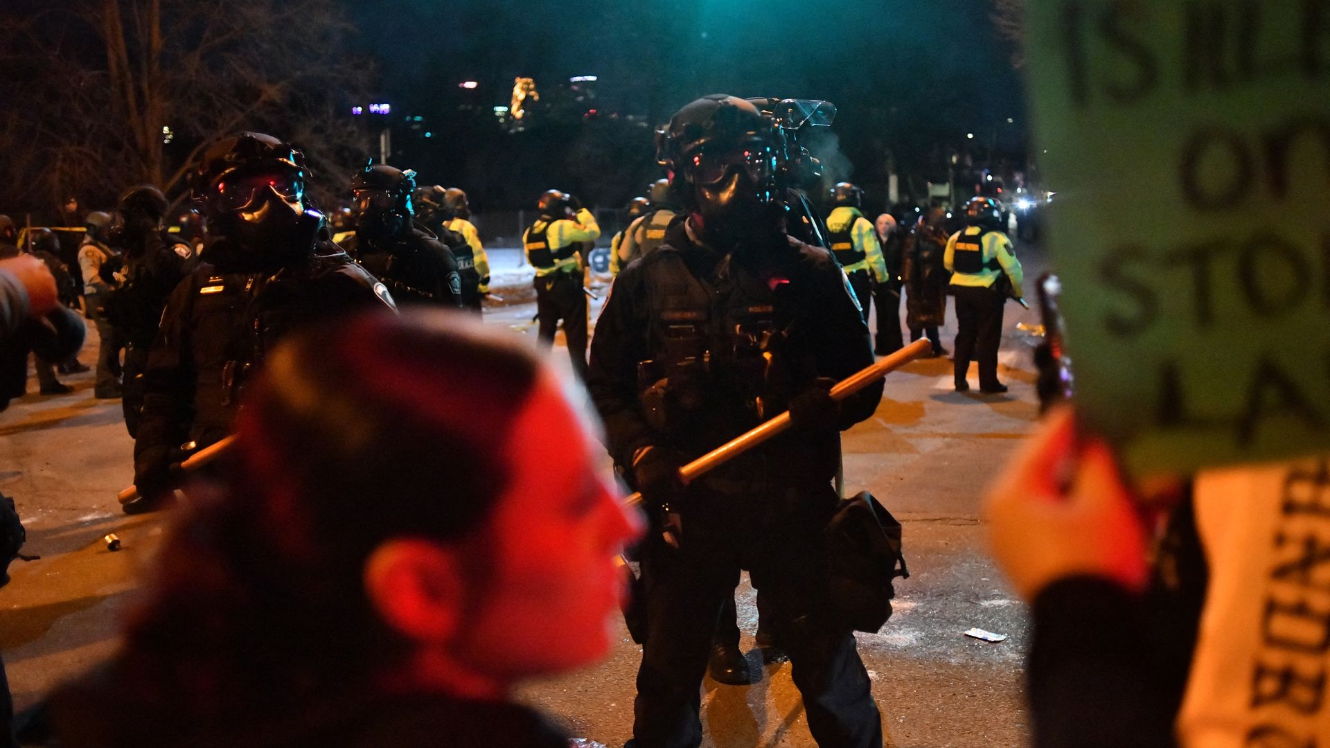 Police officers in masks and riot gear face toward protestors on a street in a nighttime scene with the Minneapolis skyline visible in the background