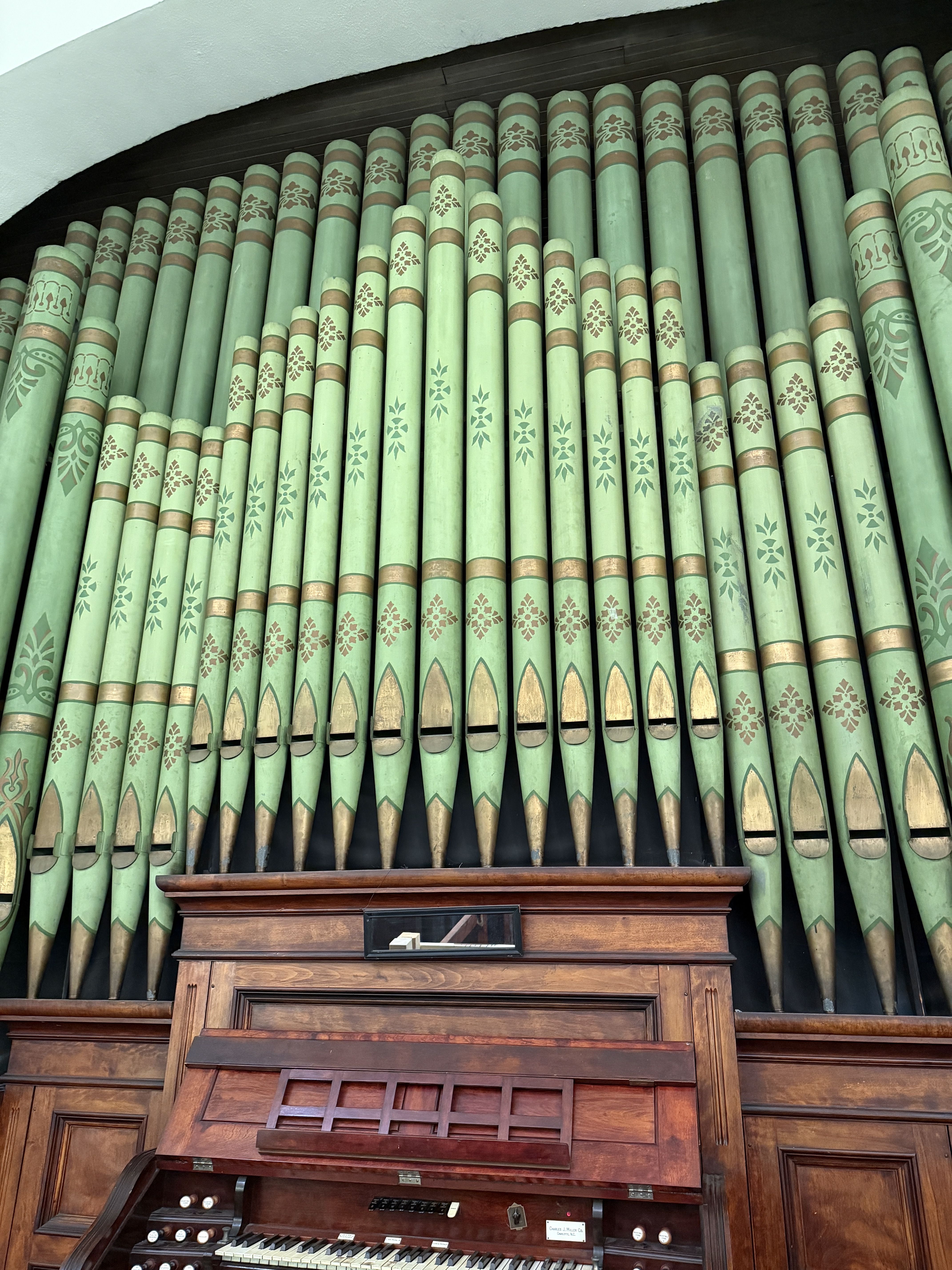 A row of tall mint-green organ pipes with gold bands and decorative floral patterns, arching above a polished wooden organ console with keys and stops.