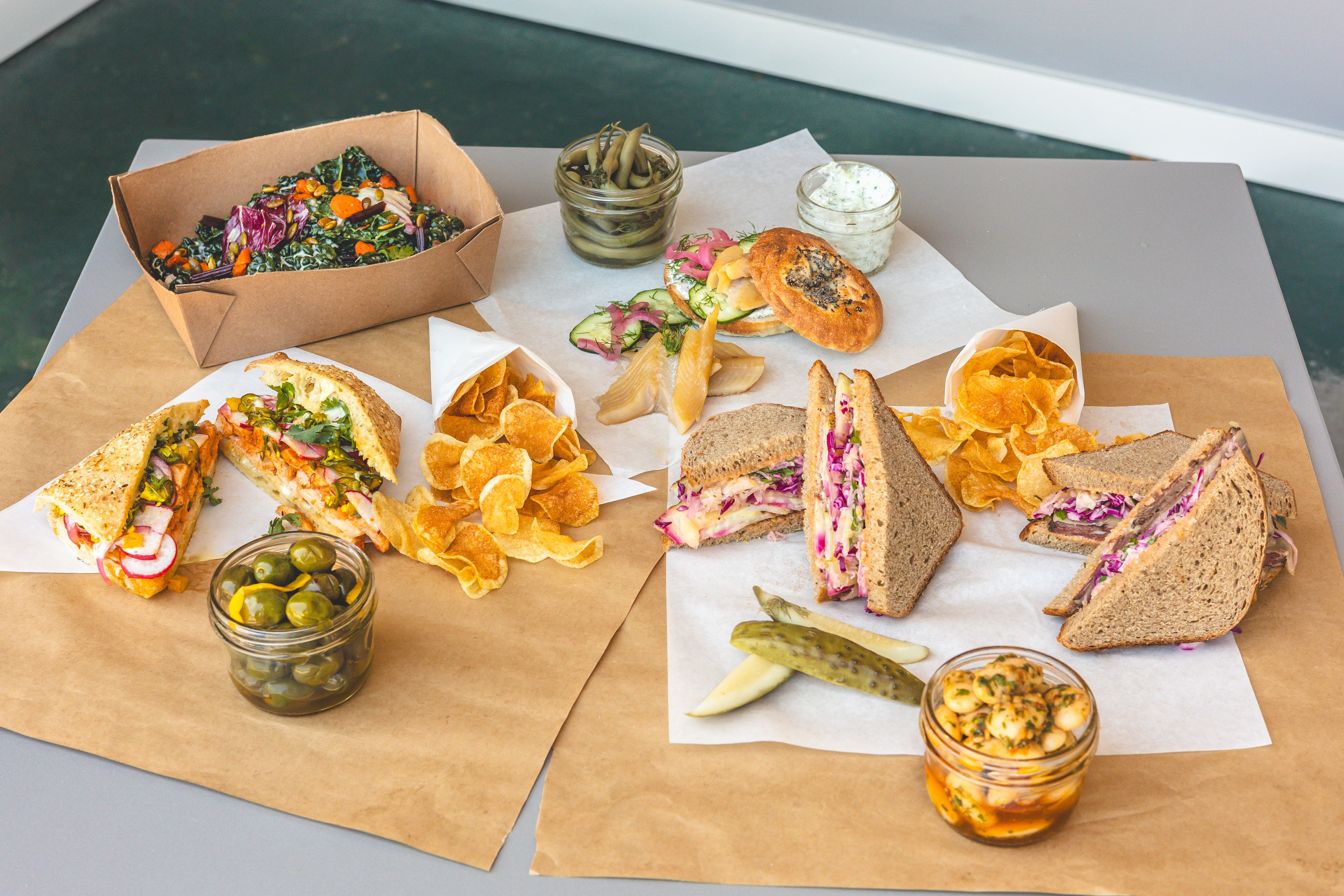Table with various sandwiches including multigrain and seeded rolls, pickles, green olives in jars, chips in white paper cones, a leafy kale salad with orange garnish, and herb dips.