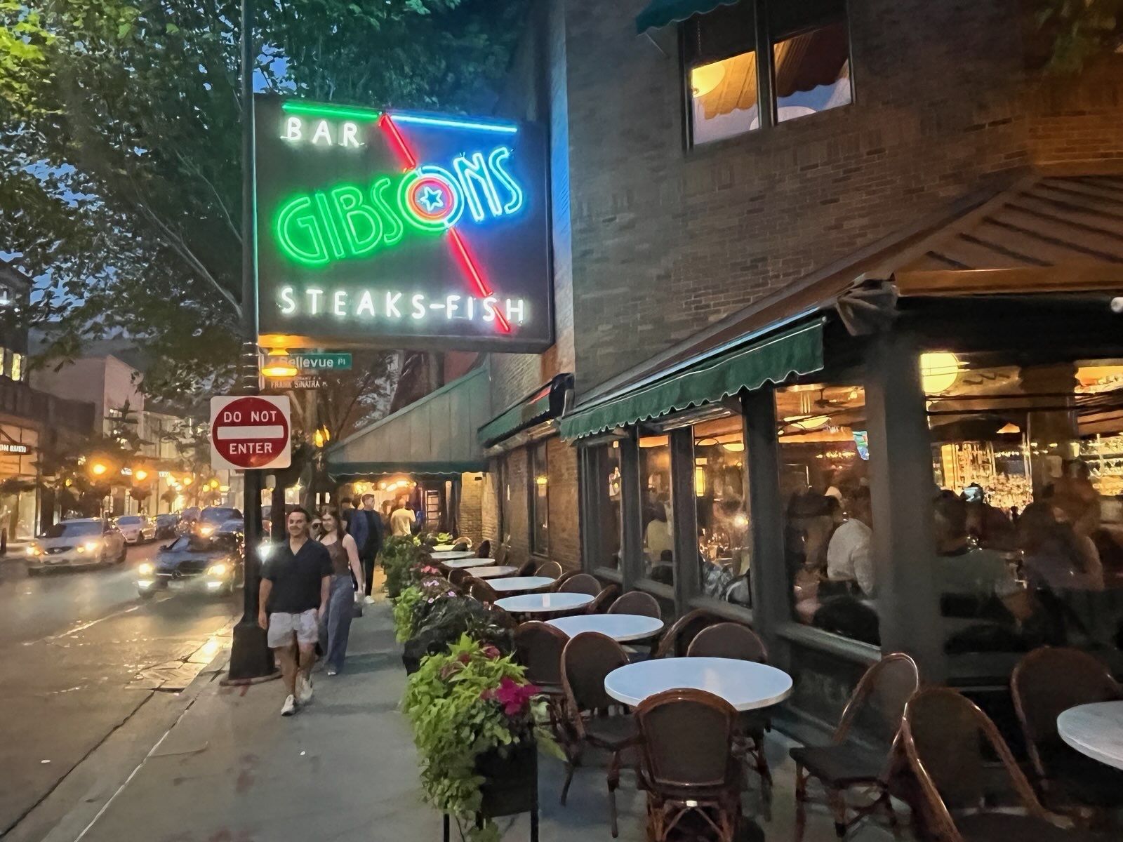Night street scene outside Gibson's Bar & Grill with a neon green/blue sign reading "GIBSON'S" and a red diagonal stripe; outdoor tables line the brick facade as pedestrians and cars pass by.