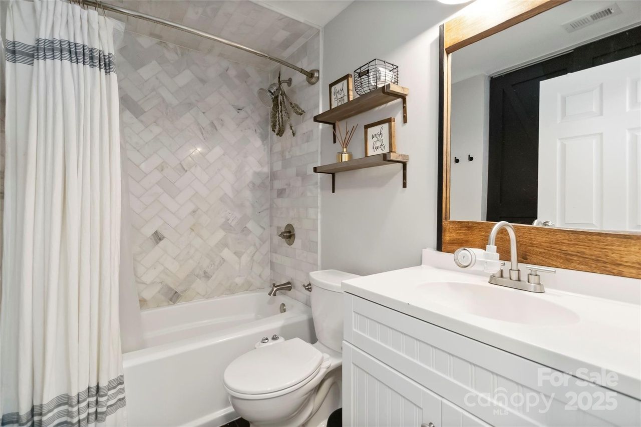 Bright bathroom with white tub, marble herringbone tile wall, white toilet, white vanity, wooden framed mirror, two wooden shelves with decor, white and grey striped shower curtain.