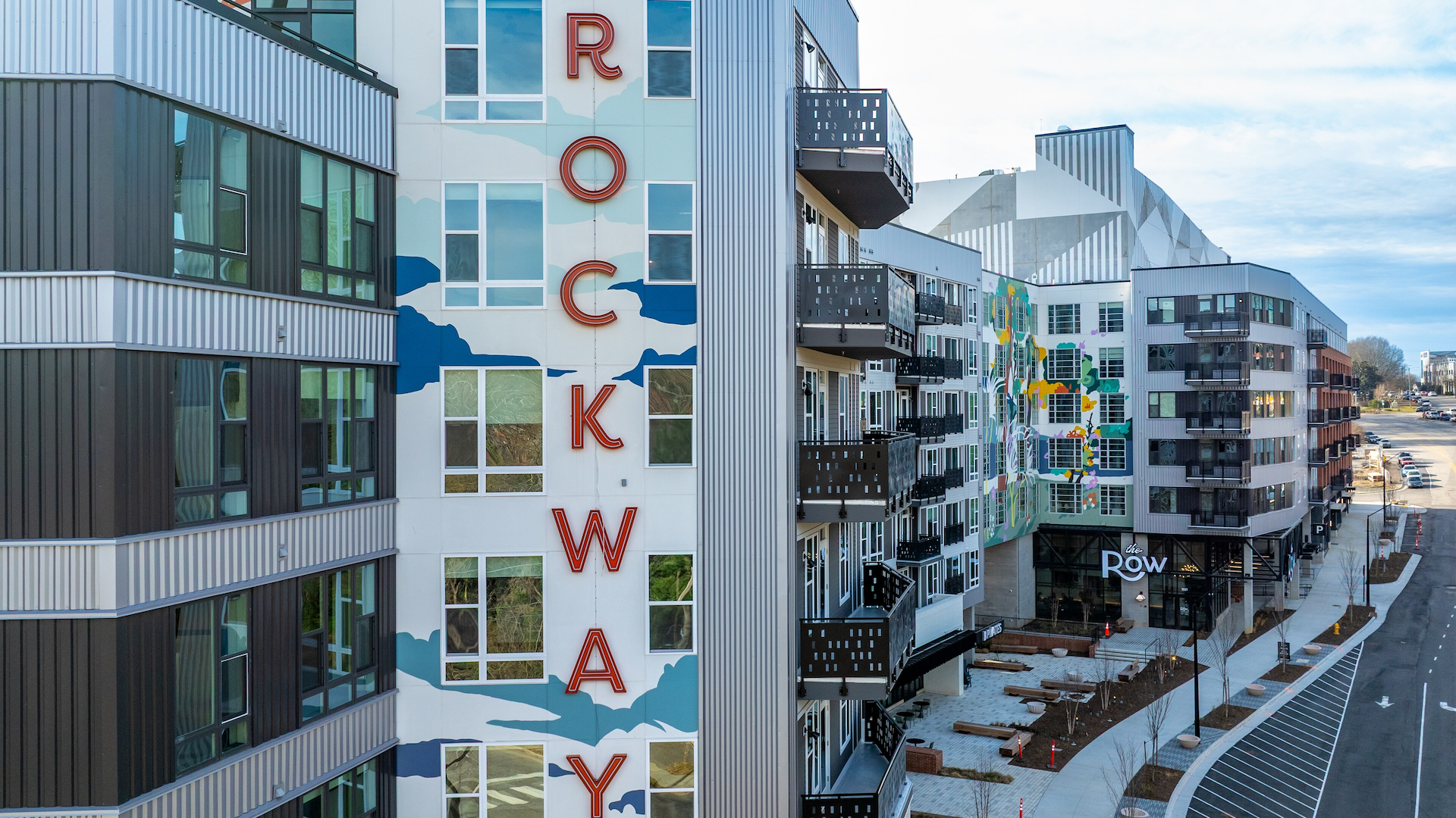 Modern multi-story apartment complex with black balconies and colorful murals, featuring the large red vertical sign "ROCKWAY" on a white wall and entrance labeled "the Row". 