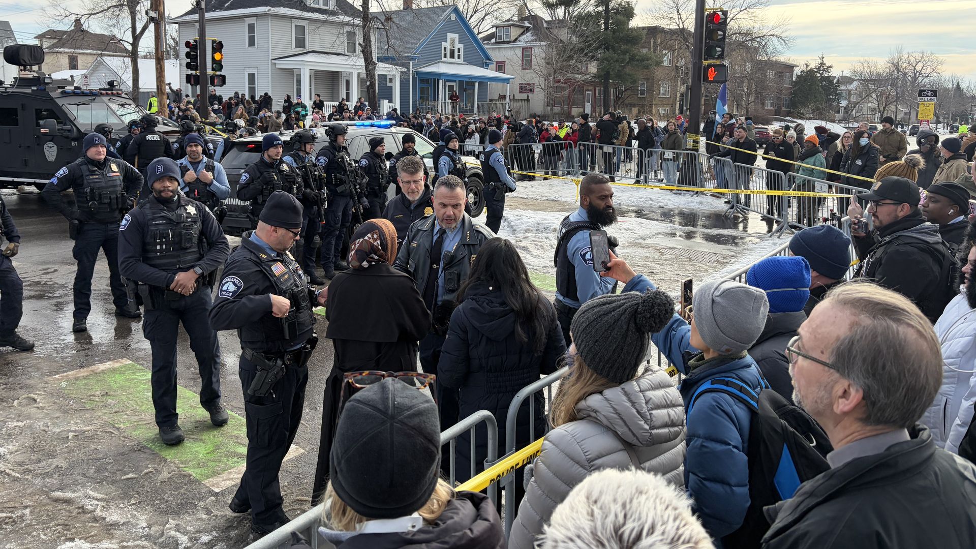 Policemen in dark uniforms monitor a crowd behind yellow police tape near an intersection with street signs reading E 34th St, with houses and winter trees in the background on a cloudy day.