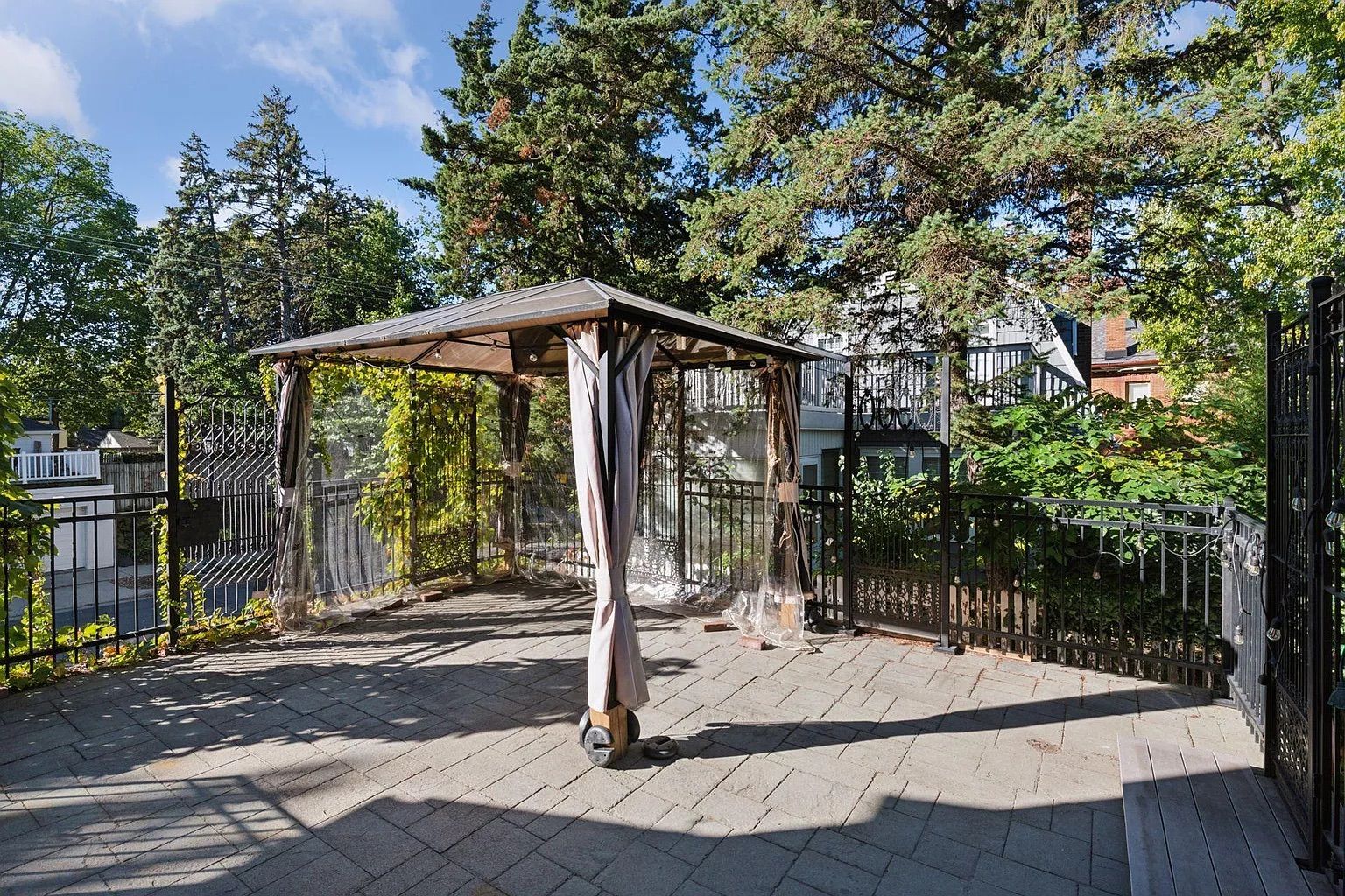 Empty outdoor patio with a metal frame canopy featuring beige curtains and clear side panels, surrounded by black metal fencing, trees, and a clear blue sky.
