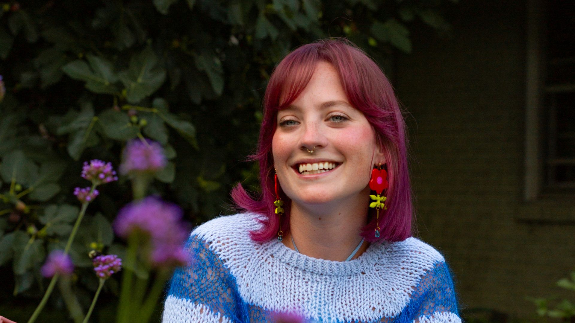 Portrait of a smiling person with pink hair, wearing a blue and white striped knit sweater, sitting on an orange metal chair in a garden. Purple flowers in the foreground, green leaves behind.