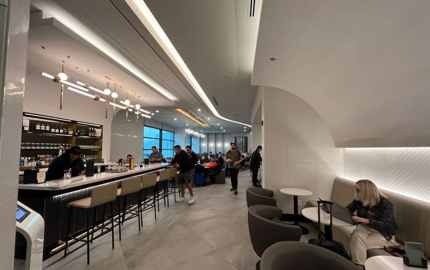 An airport lounge bar with empty counter stools and a woman sitting at a small white table with a laptop.