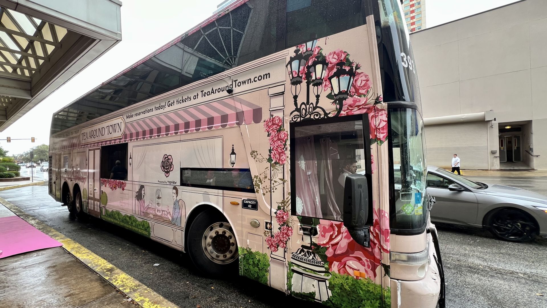 Pink double-decker bus decorated with floral and tea shop illustrations, parked on a city street on a rainy day. Text reads "Make reservations today! Get tickets at TeaAroundTown.com".