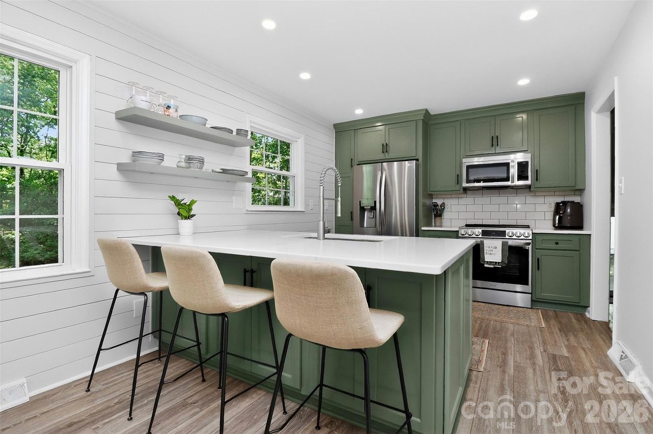 Bright kitchen with green shaker cabinets, white island and countertops, stainless steel fridge and oven, white subway tile, open shelves, beige bar stools, wood-look flooring, large windows.